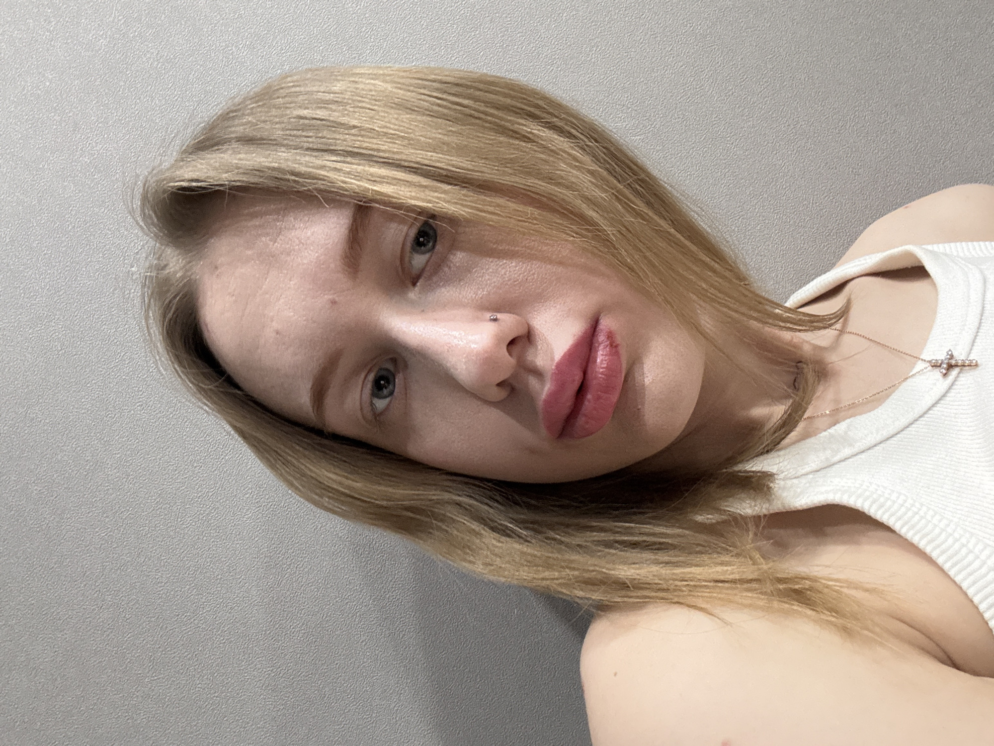 A close-up, angled portrait shot features a young woman with blonde hair, fair skin, and light-colored eyes. Her head is tilted to the right, and she is looking directly towards the camera. Her straight, blonde hair falls down around her face and shoulders.
Her face is prominent in the frame, with visible skin texture, including some minor pores or freckles on her forehead and cheek. She has full lips, colored with a glossy pink or plum-toned lipstick, and they are slightly parted. A small, sparkly piercing is visible on her right nostril.
She is wearing a white, ribbed tank top or camisole, visible on her right shoulder and chest. A delicate, fine chain necklace, appearing to be made of gold or rose gold, is around her neck. It features a small, cross-shaped pendant adorned with tiny, clear sparkling stones.
The background is a plain, light grey wall with a subtle, textured finish that is consistent throughout the visible area. The lighting appears soft and even, highlighting her features without harsh shadows. The overall impression is a casual, personal selfie.