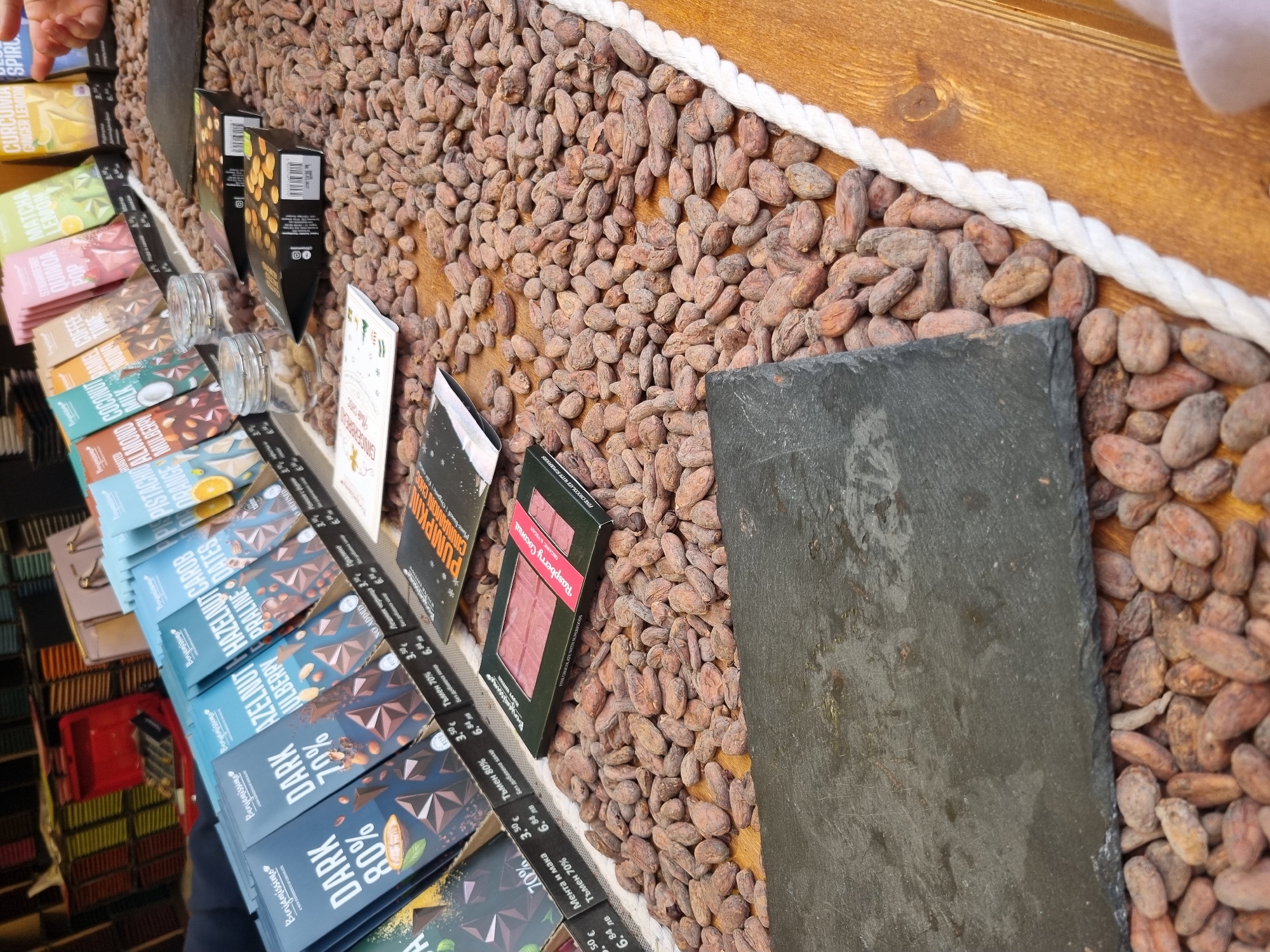 This image displays a selection of chocolate bars on display for sale, with the brand name "Benjamissimo" prominently featured. The chocolate bars are arranged in a row, likely for customers to browse and purchase. Flavors like "Dark 80%," "Dark 70%," "Hazelnut Mulberry," "Praline Dates," "Orange Mulberry," "Milk," "Strawberry Lemon," "Matcha," "Pumpkin Cinnamon Choc," "Raspberry Coconut," and "Gingerbread" are visible, indicating a variety of choices. Prices are listed in both Euros (€) and Bulgarian Lev (лв). The background features a bed of cocoa beans, adding to the aesthetic appeal of the display. A dark slate or stone surface sits amongst the beans. The items are likely displayed on a wooden surface bordered by a decorative white rope. Based on the reference to Bulgarian Lev, this could potentially be a store located in Sofia, Bulgaria. A hand appears in the upper-left corner.