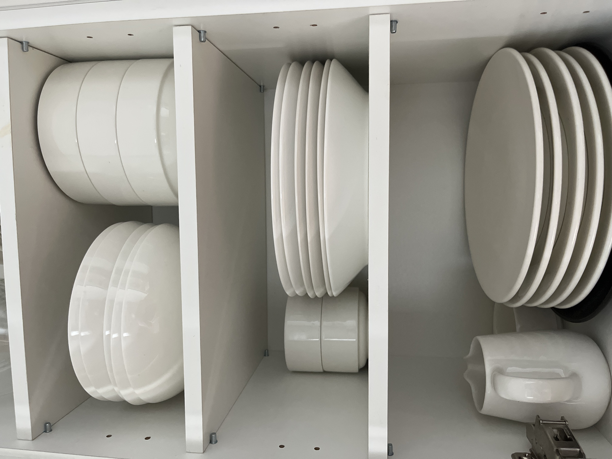 The image shows the inside of a white kitchen cabinet with multiple shelves filled with white dishes. On the top shelf, there are two stacks of white bowls, one stack of three larger bowls and another stack of four smaller bowls. On the middle shelf, there is a stack of white bowls to the left and a stack of white plates to the right. On the bottom shelf, there is a white pitcher to the left and a large stack of white plates to the right. The cabinet is clean and organized. No information about the location in Helsinki, Finland is available in the image.
