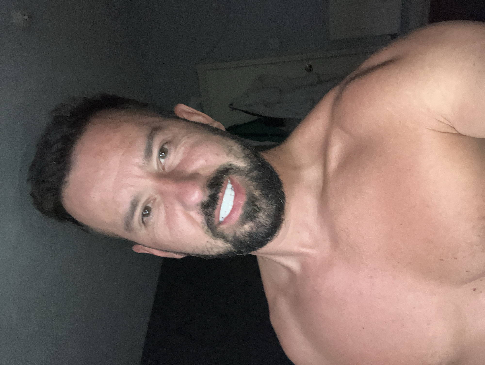A close-up, slightly rotated image of a man with short dark hair and a beard. The background is dark, with a door visible behind him. The man is shirtless, showing his upper chest and shoulder. He has light eyes and is smiling. The location is Unknown, Unknown.