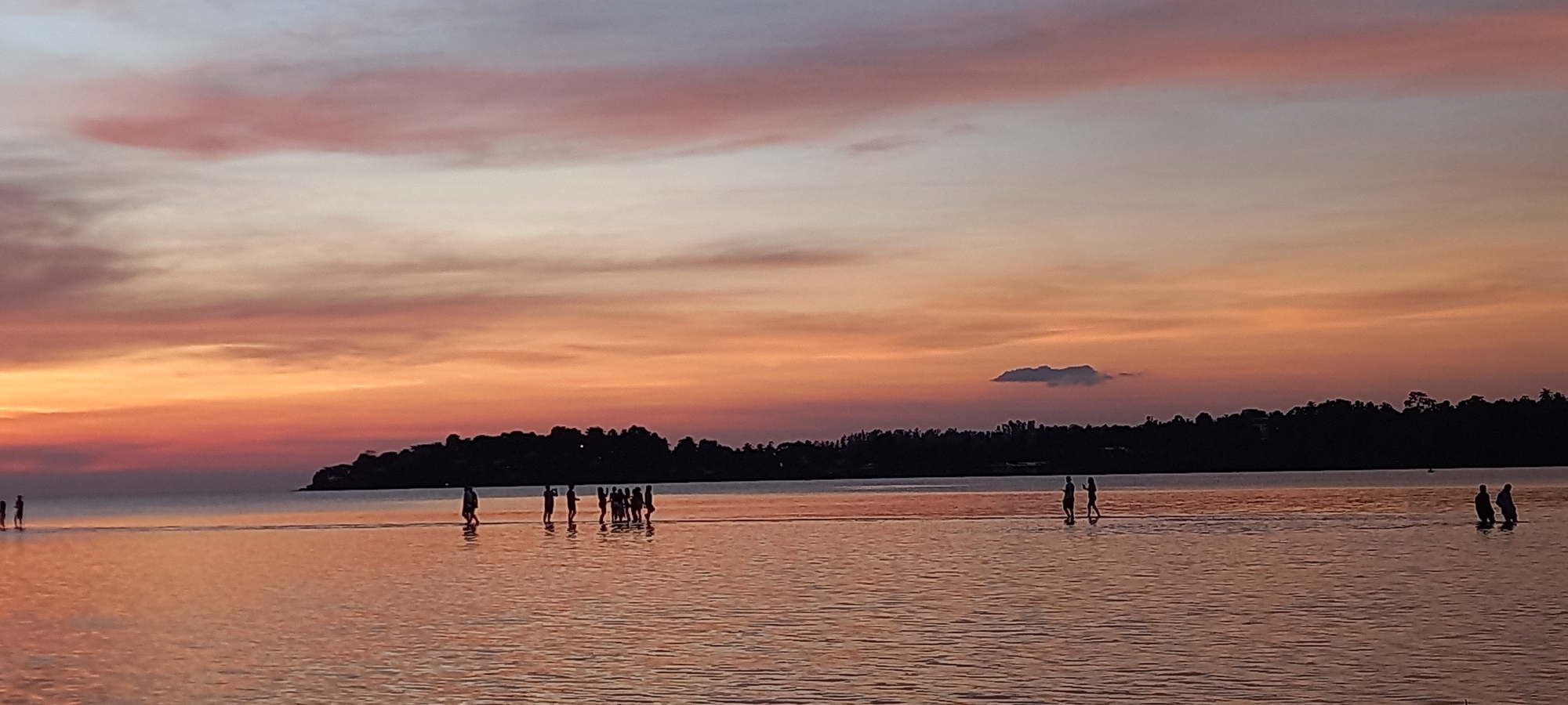 This image captures a serene sunset scene over the waters of Baan Sri Thanu, Thailand. The sky is a canvas of warm hues, transitioning from vibrant oranges and reds near the horizon to softer pinks and pale blues higher up, with delicate streaks of clouds scattered across it. A distinct, elongated cloud floats in the middle-right of the sky.
The horizon is marked by a dark silhouette of a distant landmass, densely covered in trees and foliage, suggesting an island or a forested coastline. The calm water in the foreground reflects the warm colors of the sky, creating a glowing, mirror-like surface. The water appears shallow, especially in the areas where people are standing.
Numerous individuals are visible, silhouetted against the bright sky and reflective water. On the far left, a couple of figures are wading. Towards the center, a group of approximately 6-8 people are gathered, standing close together in the shallow water. Further to the right, two figures are seen walking or standing, followed by two more figures on the far right. They appear to be enjoying the tranquil evening, perhaps walking along a sandbar or in very shallow water.
The time of day is clearly sunset, as indicated by the dramatic and colorful sky. The weather appears calm and pleasant, with no signs of disruption. The overall mood of the scene is peaceful and contemplative. There is no visible text in the image.