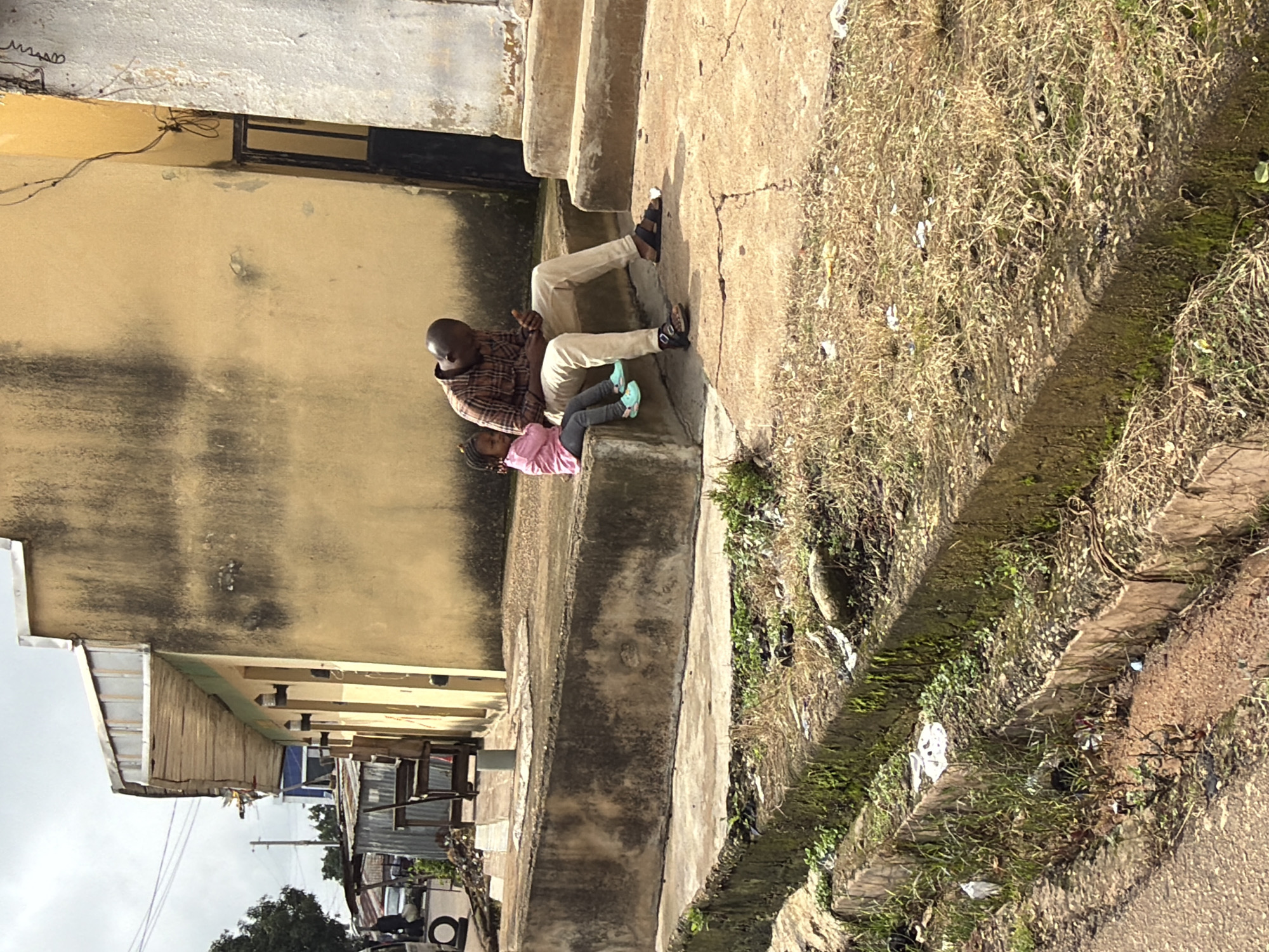 A man and a young child are sitting on a concrete ledge outside a weathered yellow building. The man, wearing a plaid shirt and light-colored pants, is looking down at his phone. The child, in a pink shirt and dark pants, sits beside him with their legs dangling. In the foreground, a concrete drainage ditch is visible, filled with dry grass, moss, and some litter. The building appears to be a commercial establishment, with a tin roofed awning extending from its side. The overall scene suggests an urban setting in Jos, Nigeria, characterized by its modest architecture and somewhat unkempt public spaces.