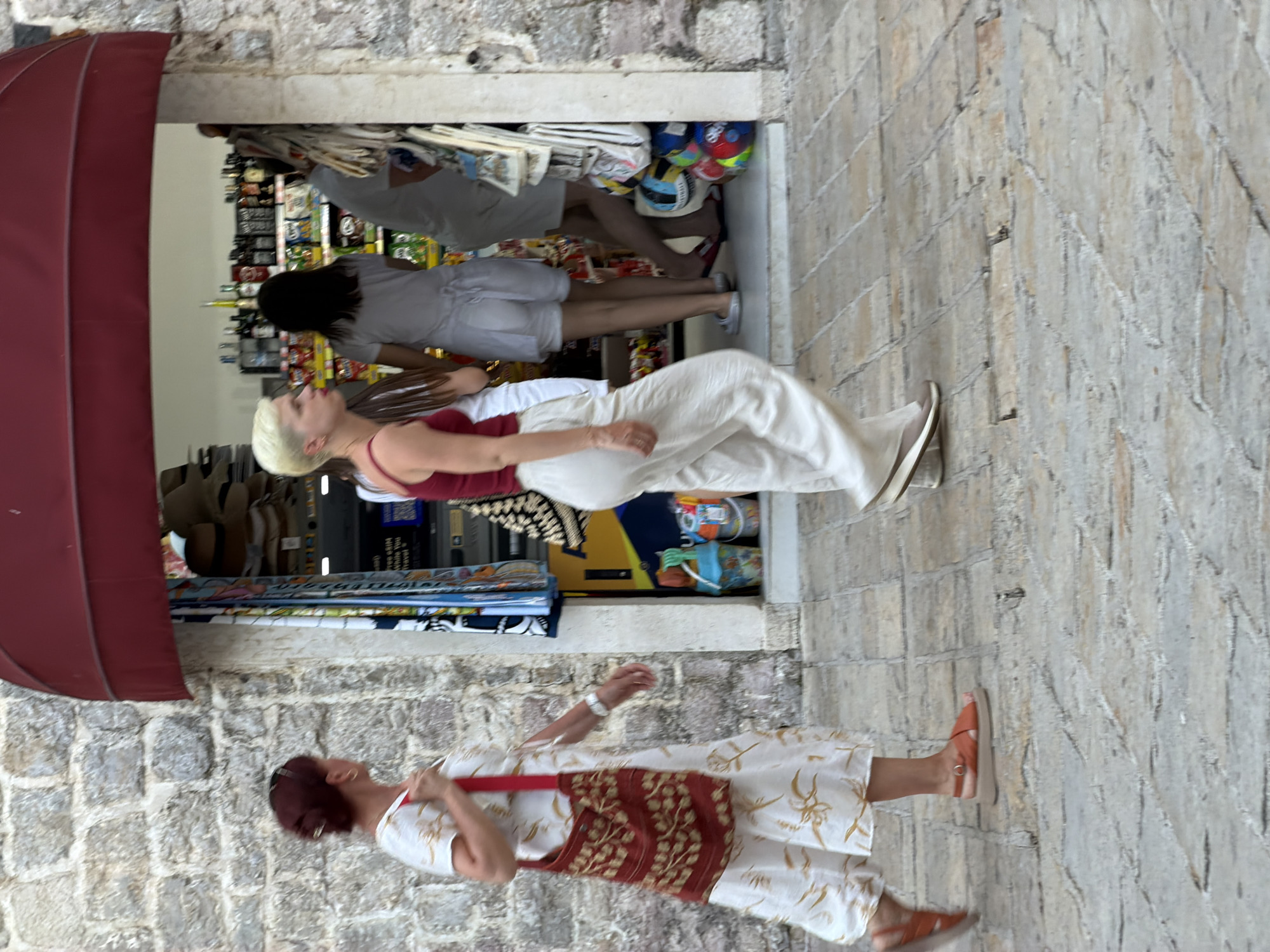 A group of people are walking past a small shop in Budva, Montenegro. The shop appears to be selling souvenirs and beach items. The building is made of stone, and there is a red awning over the shop entrance. The people in the image are dressed casually, and it appears to be daytime. The ground is paved with stone. The person in the foreground on the left is a woman with dark hair pulled back, wearing a white dress with a floral pattern and carrying a red and gold patterned bag. She is wearing orange sandals. To her right, a woman with short blonde hair is walking, wearing a red tank top and white pants. She is wearing light-colored slip-on shoes. Behind her, a person with dark hair is wearing a white t-shirt and white shorts. Further back, a person in a grey t-shirt and dark pants is also visible. Inside the shop, shelves are stocked with various items.