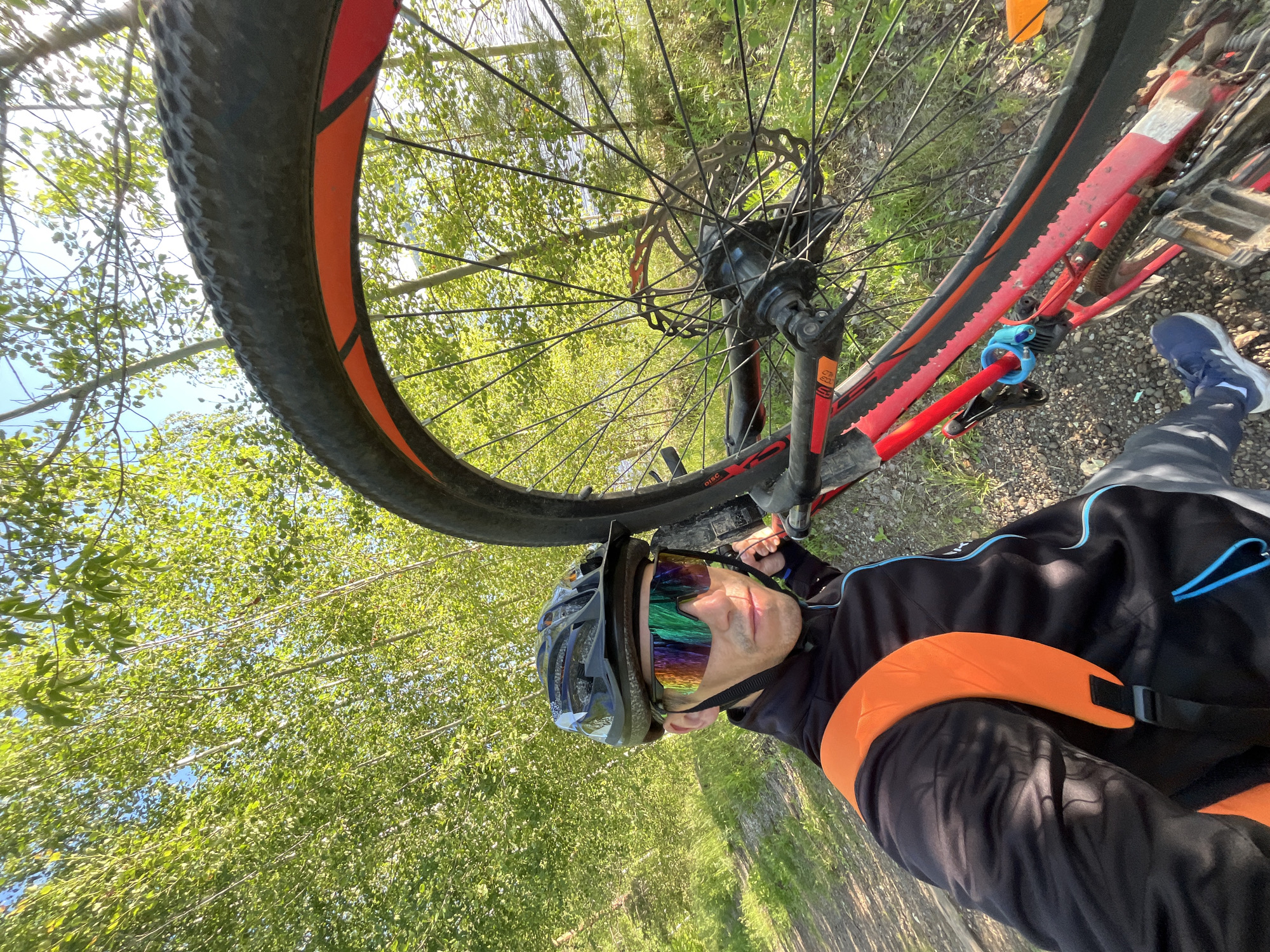 A man stands outdoors, holding a bicycle. He is wearing a black cycling jacket with blue trim, a black helmet, and colorful mirrored sunglasses. An orange backpack is on his back. The bicycle is red and black with a knobby tire. The background shows a path and abundant green trees, suggesting a natural or park-like setting. The sky is visible through the leaves, with a hint of water in the distance. The image appears to be a selfie taken by the man. The location is specified as Chaikovsky, Russia, a city situated on the Kama River, known for its industrial development and surrounding natural landscapes, often featuring wooded areas suitable for outdoor activities like cycling.