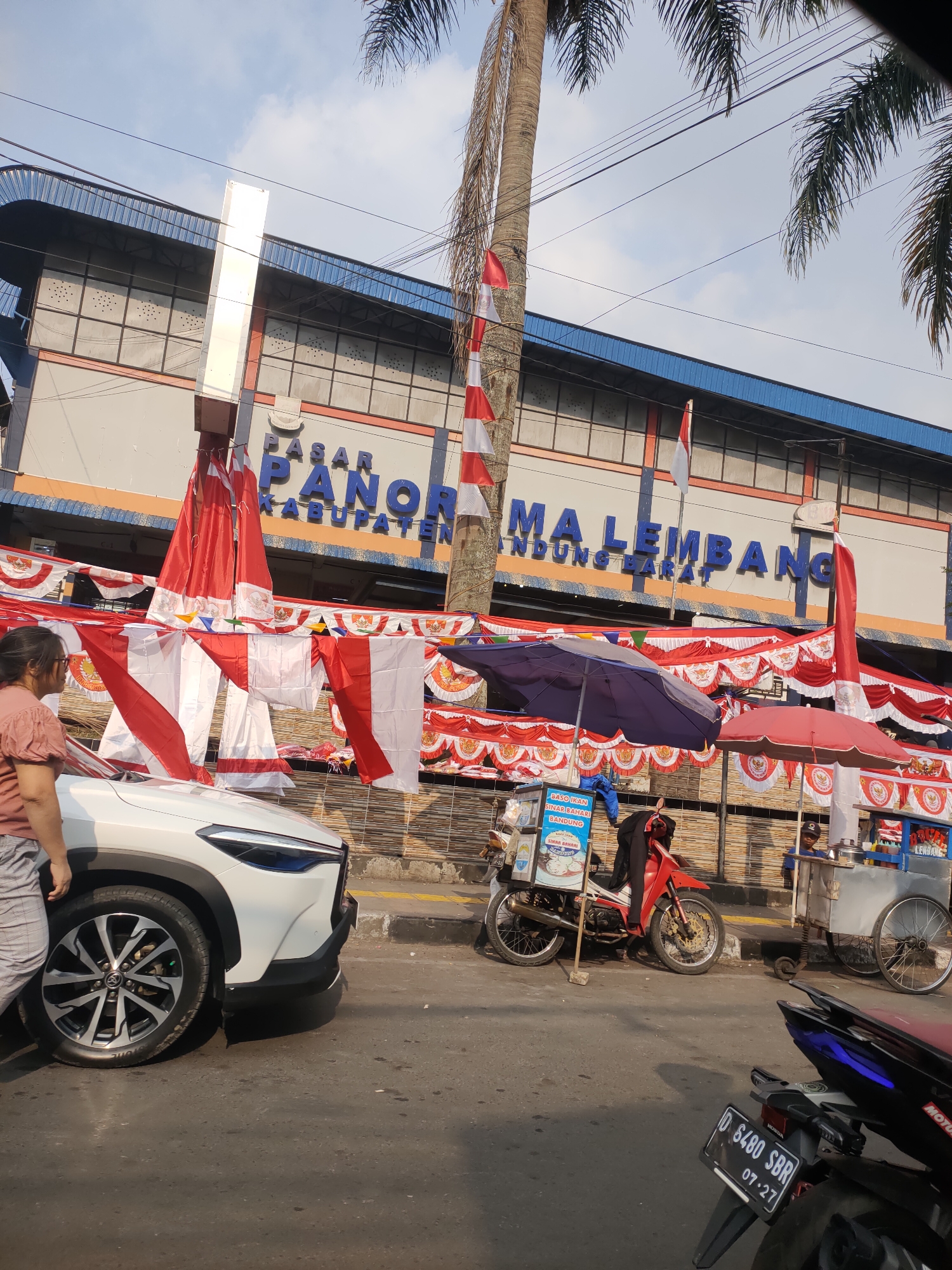 The image depicts a street scene in Lembang, Indonesia, in front of a building with "PASAR PANORAMA LEMBANG KABUPATEN BANDUNG BARAT" signage. The building appears to be a market or commercial complex. Red and white Indonesian flags are prominently displayed as decorations, hanging from poles and strung across the front of the building and along a railing. A white SUV is parked on the left side of the road, with its front end partially visible. A person wearing a light pink shirt and grey pants is standing next to the car. In the background, there are two food carts or stalls. One cart, on the left, has a blue sign advertising "BASO IKAN SINAR BAHARI BANDUNG". A red motorcycle is parked beside this cart. The other cart, on the right, is also visible with people around it. There are also two umbrellas, one dark blue and one red, providing shade over the food carts. Palm trees are visible in the upper portion of the image, suggesting a tropical or subtropical climate. The sky is clear with some clouds. The road in the foreground is asphalt. A license plate on a black motorcycle parked on the right side reads "D 6480 SBR 07-27".