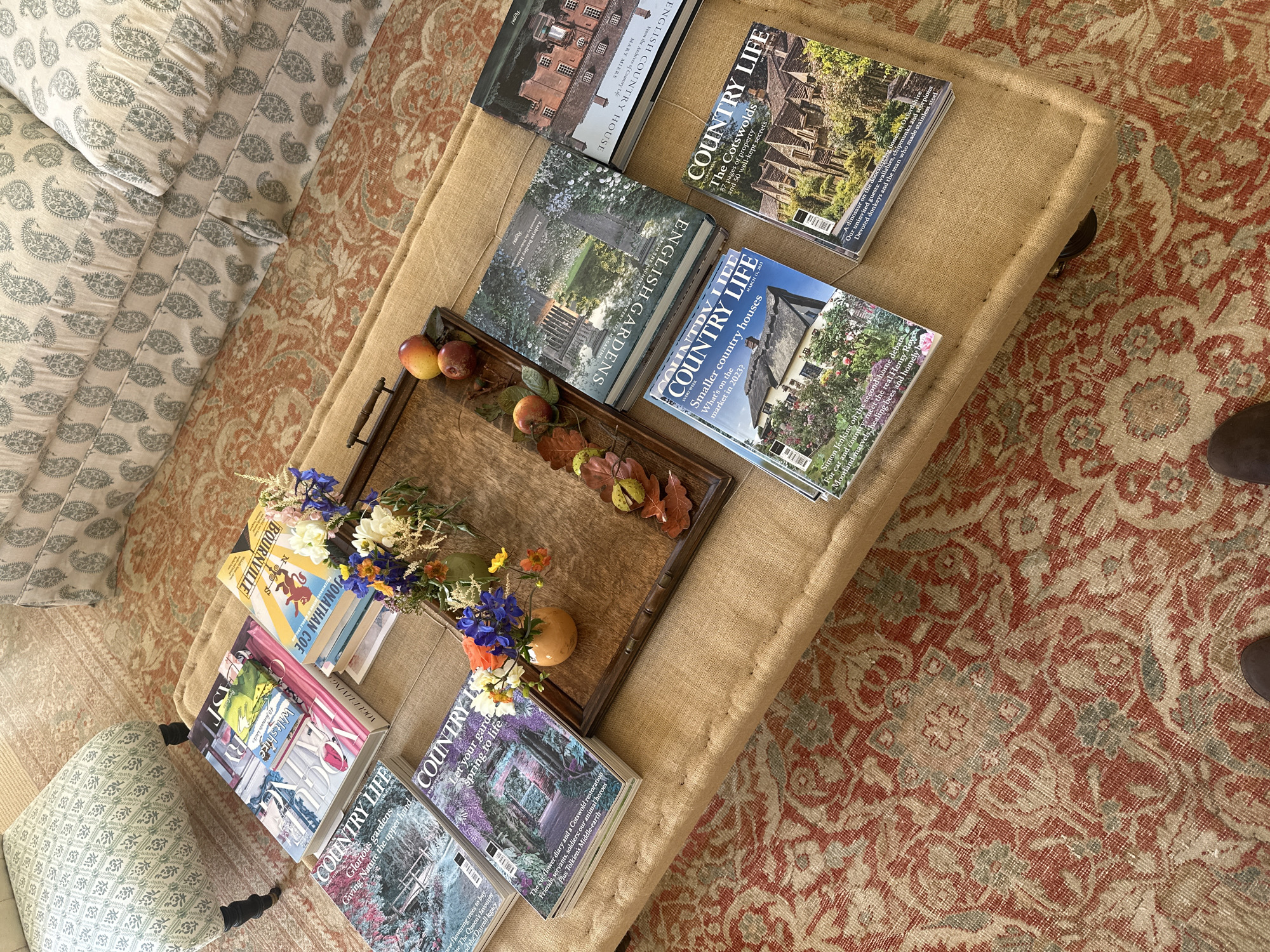Here is a description of the image:
The image shows a scene from the interior of a house in an unknown location. A burlap ottoman sits on a patterned rug. A brown, wooden tray with handles is placed on the ottoman. Within the tray are three apples, autumn leaves, and a vase with flowers. Several books and magazines are arranged around the tray on the ottoman: "Bournville" by Jonathan Coe, "Vogue Living," "Country Life," and a book called "English Gardens." A couch with a patterned design is visible in the upper left of the image. A pair of brown shoes can be seen in the lower right of the image.