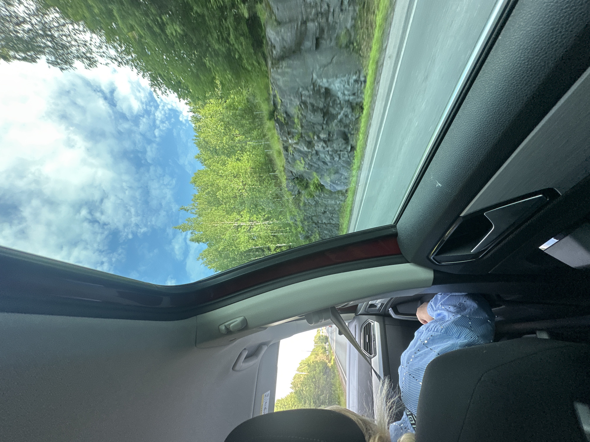 The image, taken from the interior of a moving vehicle, captures a scenic view through the passenger side window. The foreground is dominated by the interior of the car, with a black car seat visible on the left, and the door panel on the right featuring a metallic handle. A person, partially visible, is seated in the passenger seat, wearing a blue and white striped shirt with silver embellishments. Their head is turned away from the camera, and their arm is resting on the armrest.
The window reveals a road stretching into the distance, with a lush green forest and a rocky incline flanking the right side of the road. The sky is a vibrant blue, dotted with fluffy white clouds, indicating a clear day. The motion blur in the image suggests the vehicle is in transit. The environment appears to be a rural or semi-rural area, characterized by natural scenery rather than urban infrastructure.