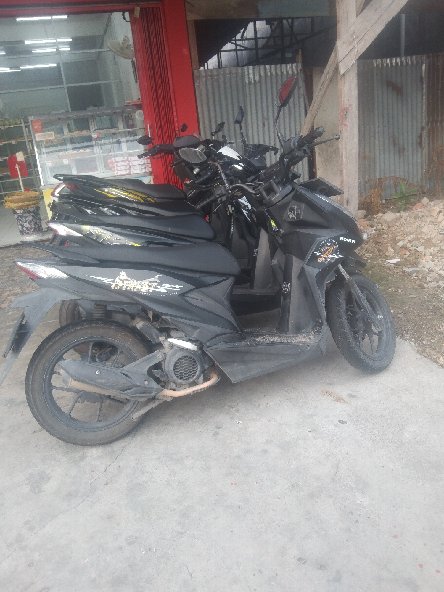 This image captures a row of motorcycles parked in front of a shop in Siak Sri Indrapura, Indonesia. The motorcycles, predominantly black and dark-colored, are the main focus. The closest motorcycle in the foreground has the word "Street" and "Beat" prominently displayed on its side panel, along with a "Honda" logo. Other motorcycles are parked behind it, forming a line.
The environment suggests an outdoor commercial area. To the left, a portion of a shop is visible, with red corrugated metal doors and a display case inside that appears to contain food items, possibly pastries or bread. Overhead fluorescent lights are visible inside the shop. To the right, there is a rustic wooden structure and a corrugated metal wall, indicating an unfinished or industrial-looking part of the building or surrounding area. The ground is paved with concrete, which is cracked in places, and debris such as small stones and dry leaves are scattered around. The overall impression is a moderately urban or semi-urban commercial setting.