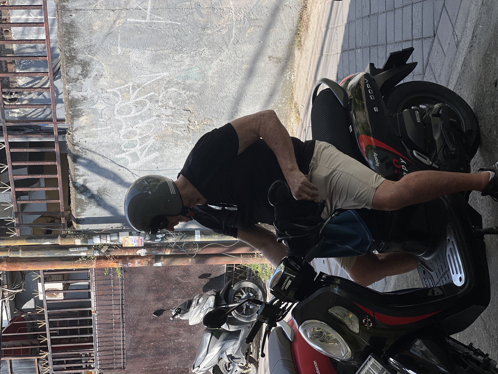 The image depicts a man wearing a black t-shirt, khaki shorts, and a green helmet, sitting on a black scooter in Kuta, Indonesia. The scooter has red and black accents and the word "SPORTY" is visible on its side. Another scooter is parked behind him, and a wall with graffiti is in the background. The scene appears to be outdoors on a sunny day, with the ground paved with square tiles. The man's hands are on the scooter's handlebars.