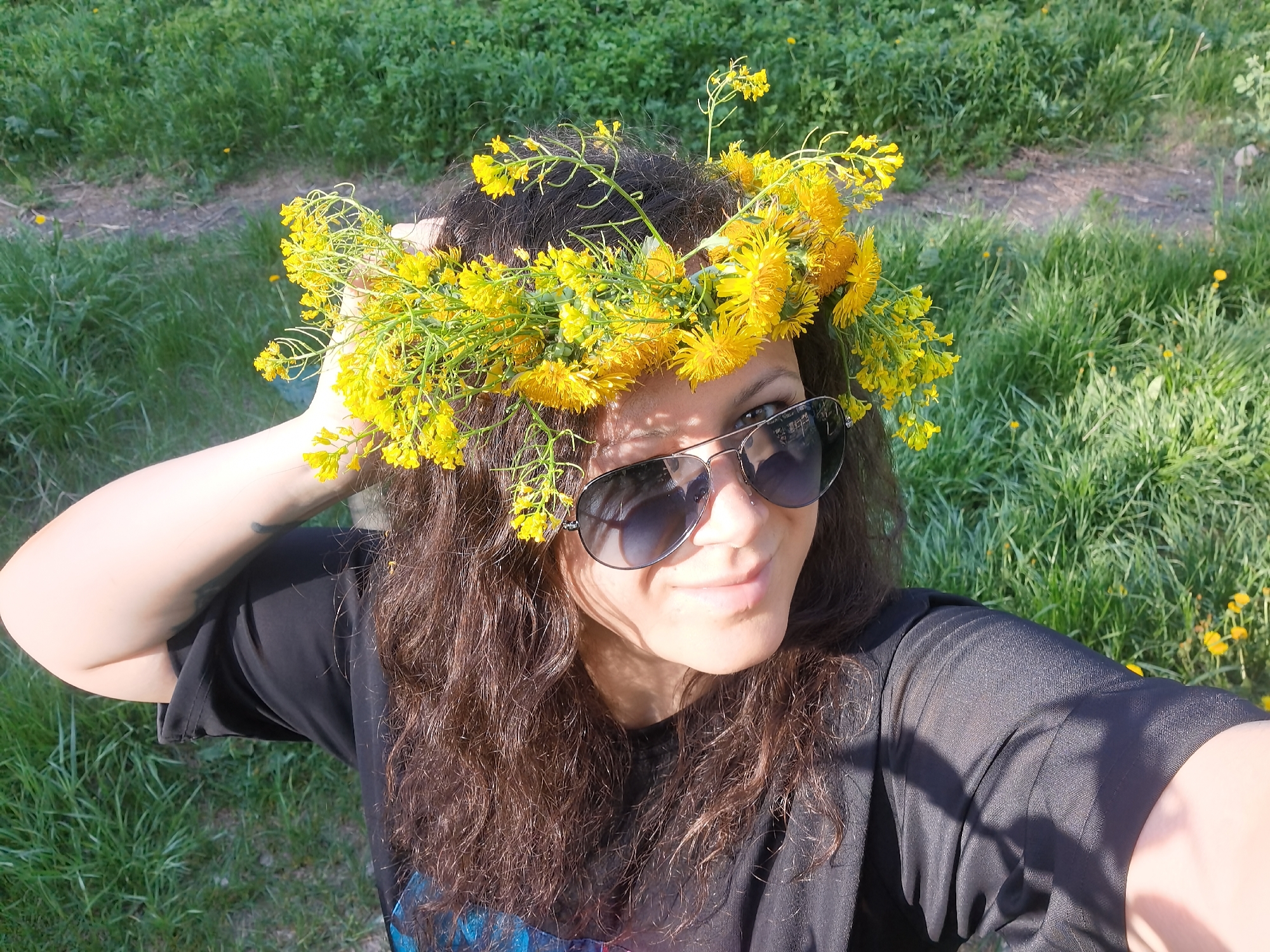 A medium, eye-level shot captures a person in Perm, Russia. The person is centered, adorned with a crown of yellow wildflowers, likely dandelions, and wearing aviator sunglasses, a black t-shirt and appears to be taking a selfie with the green grass and wildflowers in the background, some yellow weeds are visible in the grass. The person is touching the wildflower crown in her hair.