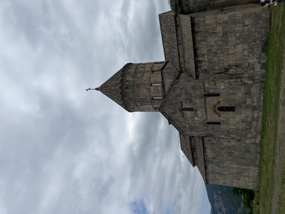 The image displays the Tatev Monastery, situated in Tatev, Syunik Province, Armenia. The scene features the monastery's main church, constructed from light to dark gray cut stone blocks. A prominent circular drum, topped by a conical roof with a small dark cross at its apex, rises from the central structure. The drum features several narrow, vertical rectangular windows. Below the drum, gabled roof sections extend, supported by thick stone walls with narrow, arched windows. The roof tiles appear to be made of dark, possibly weathered, stone.
In the foreground, a patch of uneven green grass occupies the bottom portion of the frame. The background on the left reveals distant, undulating mountains or hills with some green foliage. The sky is largely overcast, dominated by varying shades of grey clouds, with a small patch of lighter sky visible on the far left.
At the bottom right corner of the frame, two individuals are partially visible. One person wears a white top and dark pants, facing away from the camera, while the other is mostly obscured. They are standing near the base of the monastery wall. No specific actions or interactions are discernible.