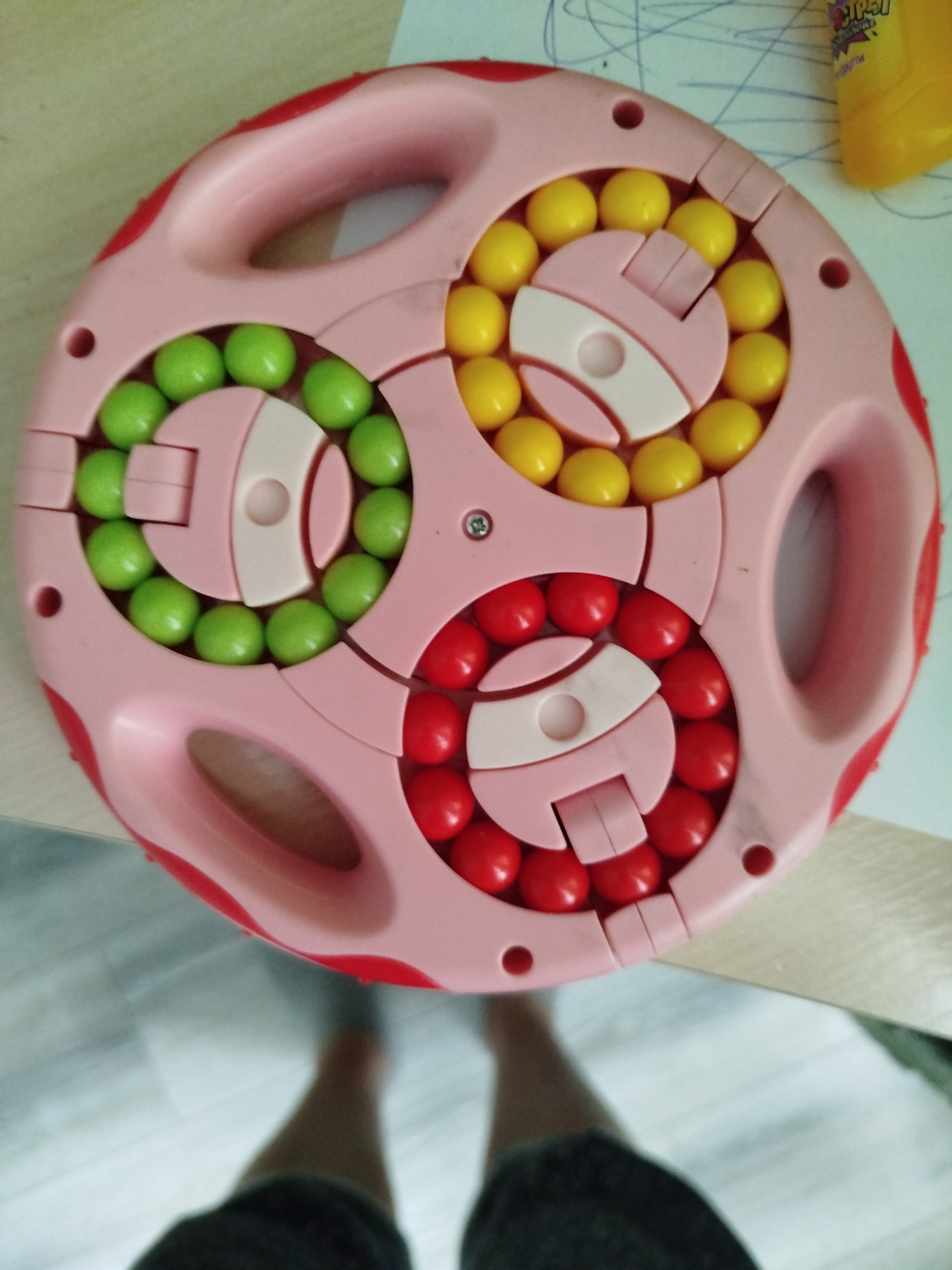 The image shows a pink puzzle toy filled with red, green, and yellow beads. The toy has three circular sections, each containing beads of a single color. The puzzle is placed on a wooden surface, and the legs of a person are visible in the foreground, suggesting they are sitting or standing near the toy. The background is blurred but appears to be an indoor setting. The toy is a common type of fidget or educational toy, and its presence does not provide any specific information about the location within Крывасельскі сельскі Савет, Belarus.