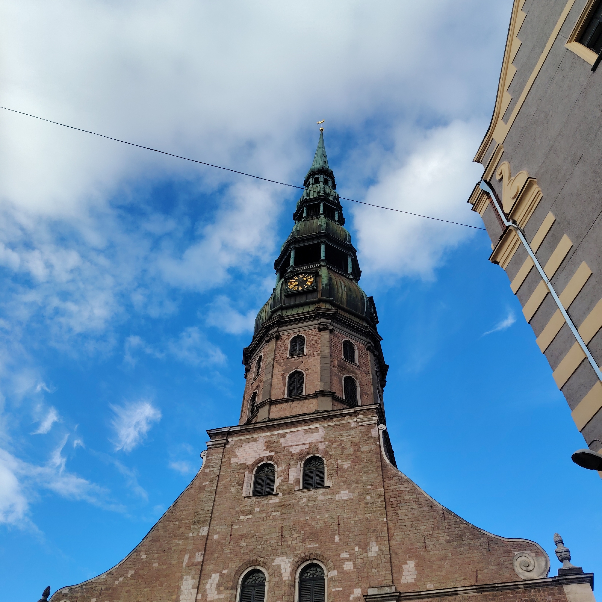 The image shows a low-angle view of the tower of St. Peter's Church in Riga, Latvia, against a bright blue sky with scattered clouds. The tower, constructed of red brick and stone, features a tall, slender spire topped with a gold rooster weather vane. A prominent clock face with gold Roman numerals is visible on one level of the tower. To the right, a portion of a grey building with yellow decorative accents and a steeply pitched roof is visible, framing the church tower. A thin wire stretches across the sky in the upper left quadrant of the image.