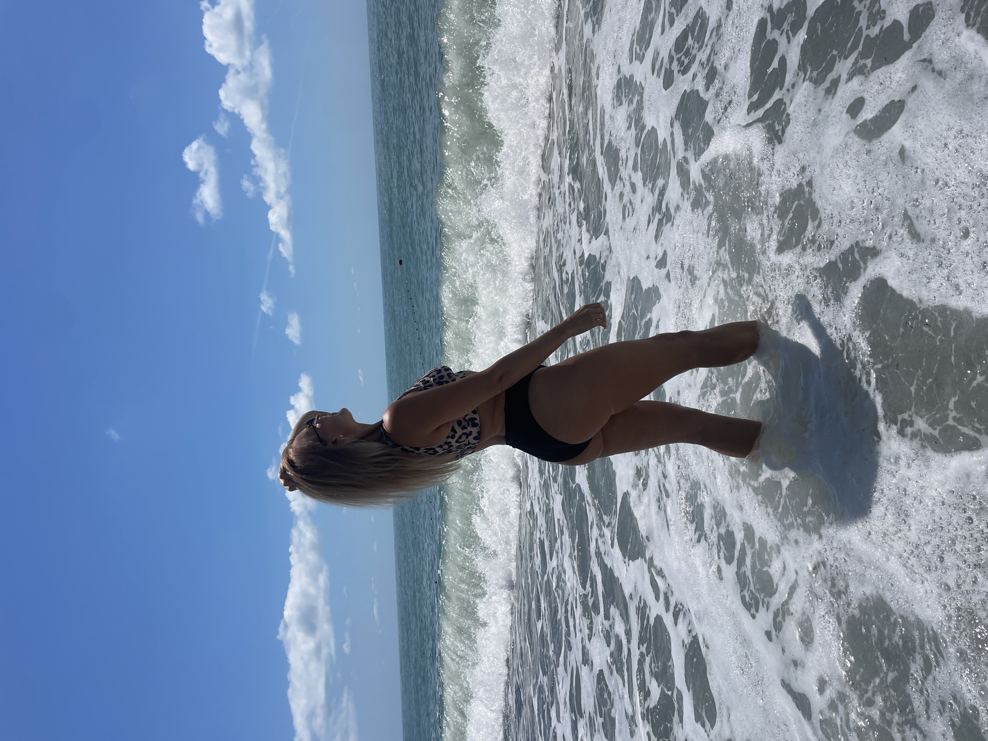 A woman stands in the shallow ocean water, facing away from the camera and to the right. She has blonde hair pulled back and is wearing a leopard print bikini top and black bikini bottoms. The ocean is rough with a large wave breaking behind her. The sky is blue with scattered white clouds. The scene is located on a beach in Sochi, Russia.