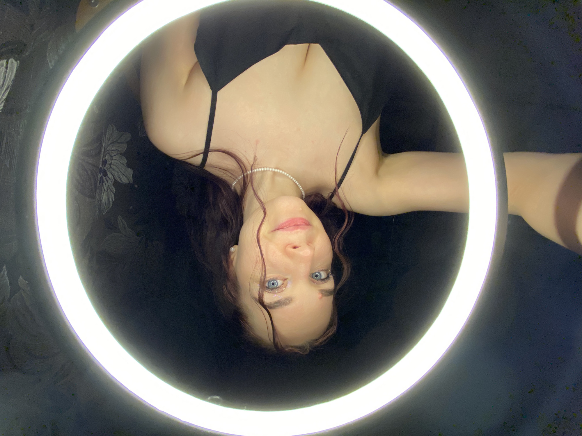 A woman with blue eyes, brown hair, and fair skin is centrally framed within a bright white ring light. She is wearing a black top with thin straps and a pearl necklace. Her face is slightly tilted upwards. The background is dark, with a pattern of floral designs visible on the left side. A portion of an arm is visible on the right. The location is not discernible from the image alone.