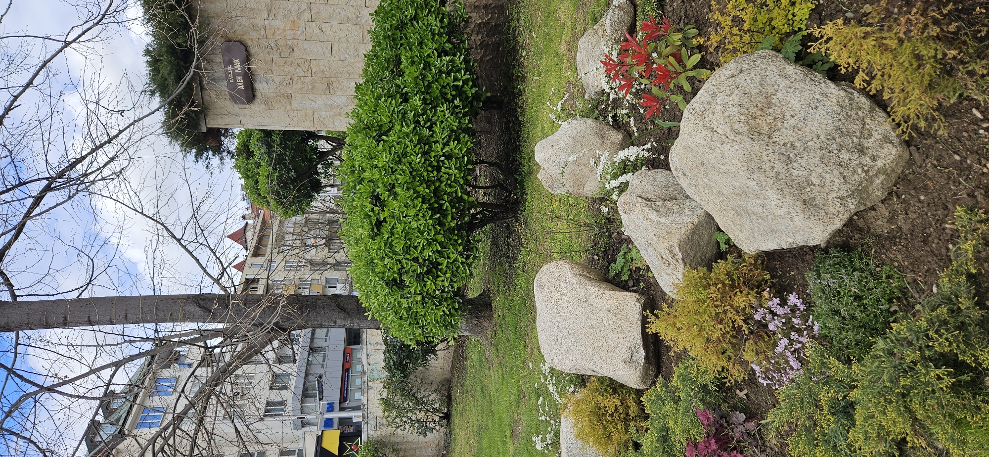 The image shows a section of a park or garden in Blagoevgrad, Bulgaria. The view is ground-level and angled, capturing a curated landscape featuring rounded bushes, large, rough-textured rocks, and a variety of colorful flowering plants. The vegetation is dense and appears well-maintained. In the background, a multi-story building is visible. The sky is partly cloudy. A tree is in the foreground. A sign is posted on a nearby wall.