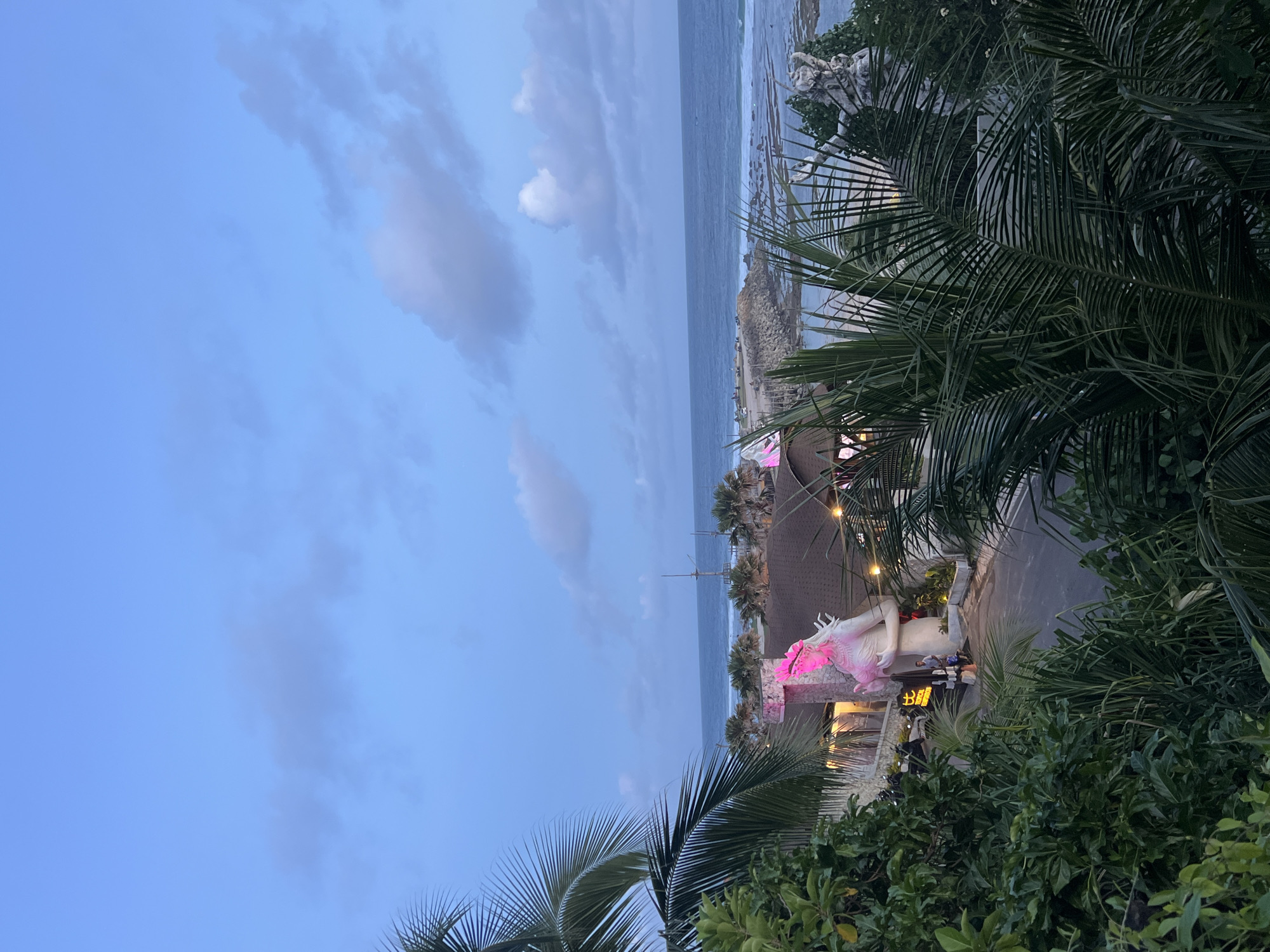 The image is taken from an elevated position, overlooking an outdoor scene in Kuta Selatan, Indonesia. The sky is a gradient of blue hues, transitioning from a lighter shade near the horizon to a deeper blue above. Soft, scattered clouds are visible in the sky. The ocean is a muted blue-gray, with waves breaking near the shoreline.
In the midground, palm trees and other tropical foliage frame the view. A building with a dark roof and string lights is present. A large, white statue with pink lighting accents is positioned near the building's entrance. People are standing and interacting in front of the building. The location is along a road, and vehicles are parked nearby. The scene captures a tropical setting at dusk.