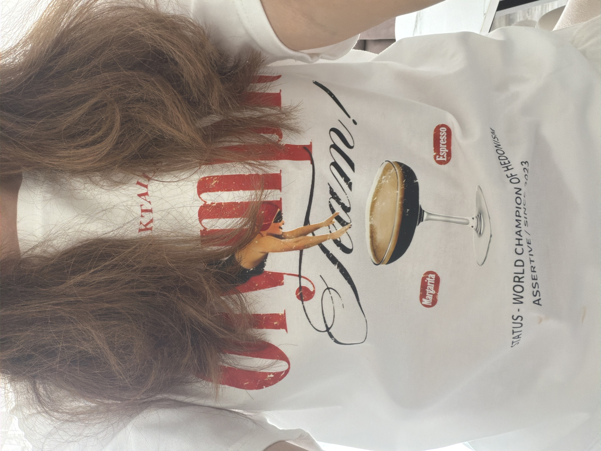 The image shows a close-up of a person wearing a white t-shirt with a graphic design. The design features large red letters that appear to spell out "ON THE..." followed by a stylized script that reads "Team!". A graphic of a person in a diving pose is superimposed on the letters. Below this, there is an image of a cocktail glass filled with a dark liquid, with the labels "Margarita" and "Espresso" on either side. At the bottom of the t-shirt, there is text that reads "STATUS - WORLD CHAMPION OF HEDONISM ASSERTIVE/SINCE 2023". The person's brown hair falls over the t-shirt. The background is out of focus and appears to be an indoor setting. There is no indication of the city of Malinovka, Russia, or any other specific location.