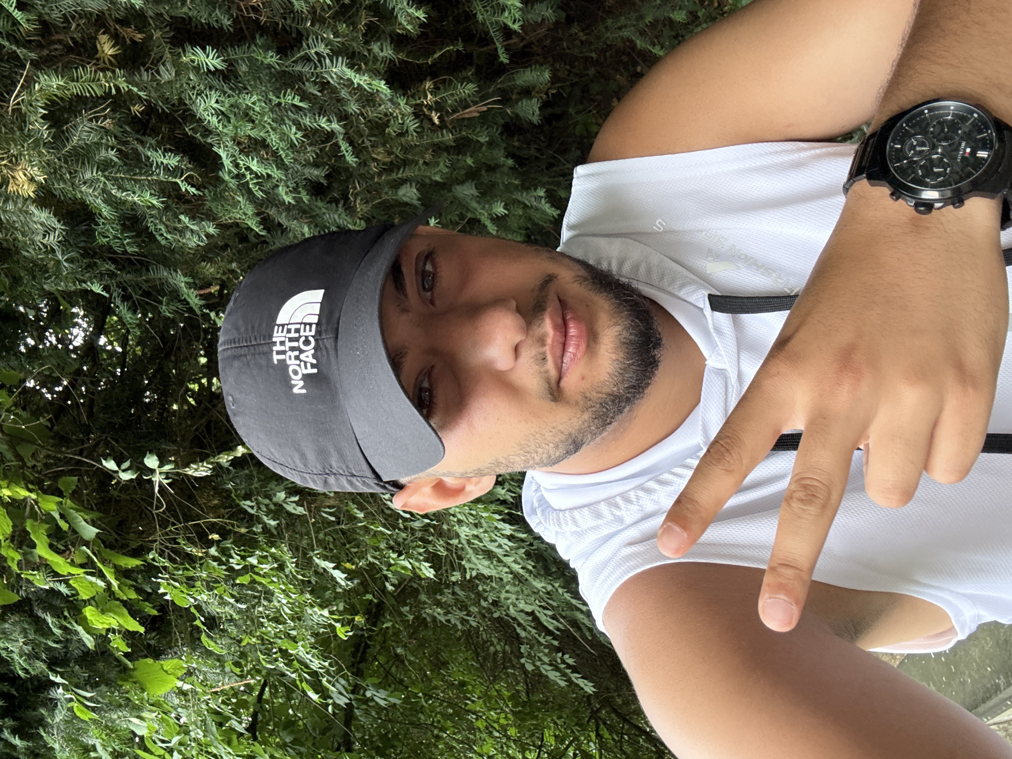 A man is pictured from the chest up, wearing a black "The North Face" baseball cap, a white sleeveless athletic shirt, and a black watch. He is giving a "peace sign" with his right hand. He has a short, dark beard and appears to be of Middle Eastern descent. The background consists of dense green foliage suggesting a park or wooded area. The location cannot be determined as Liestal without additional identifying details.