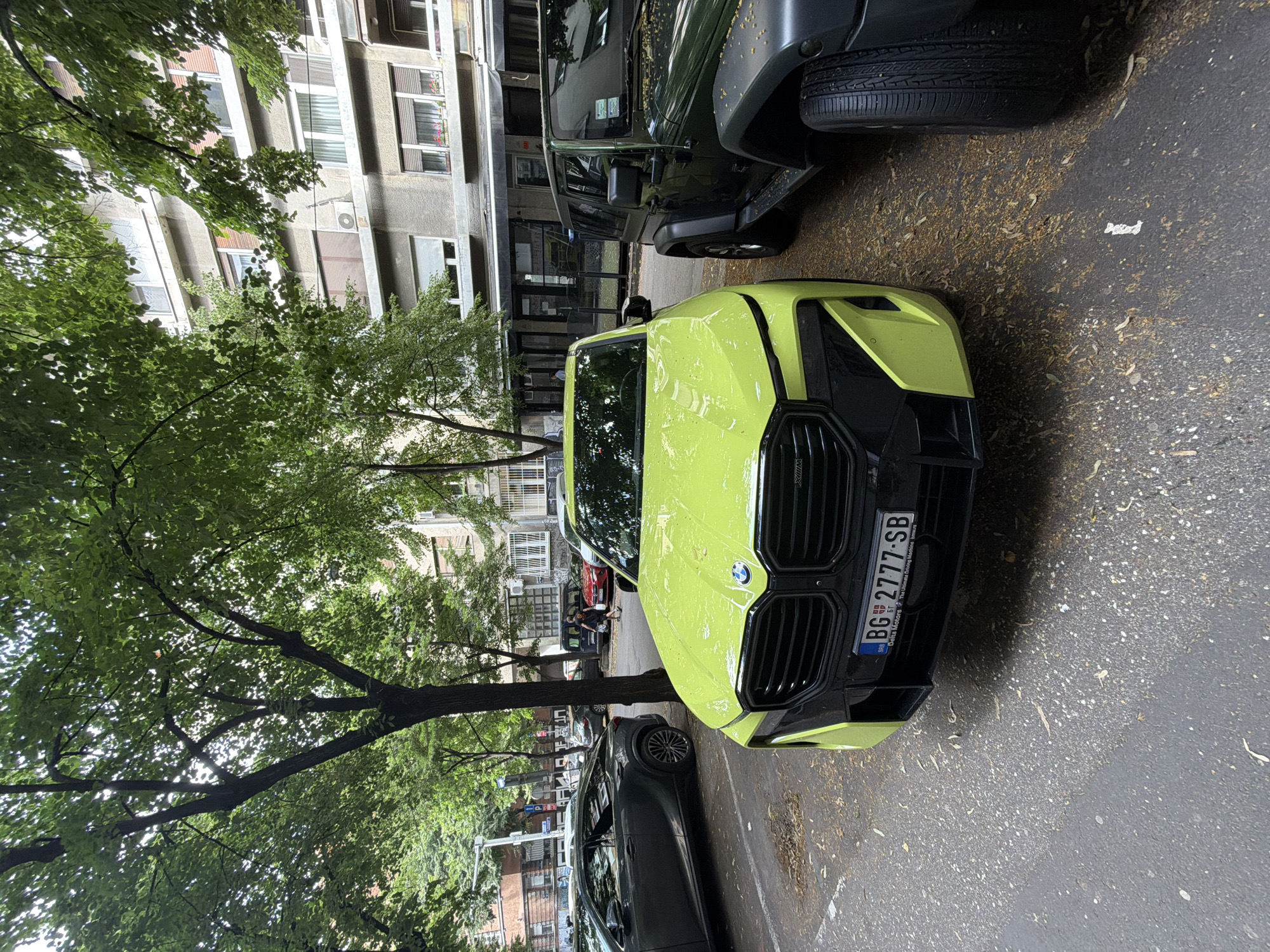 A bright lime green BMW XM is parked on a street in Belgrade, Serbia, with a dark green Jeep parked next to it. The BMW has a license plate that reads "BG 2777 SB". The street is lined with trees that are shedding leaves, and a multi-story building with balconies and windows is visible in the background. The sky is overcast.