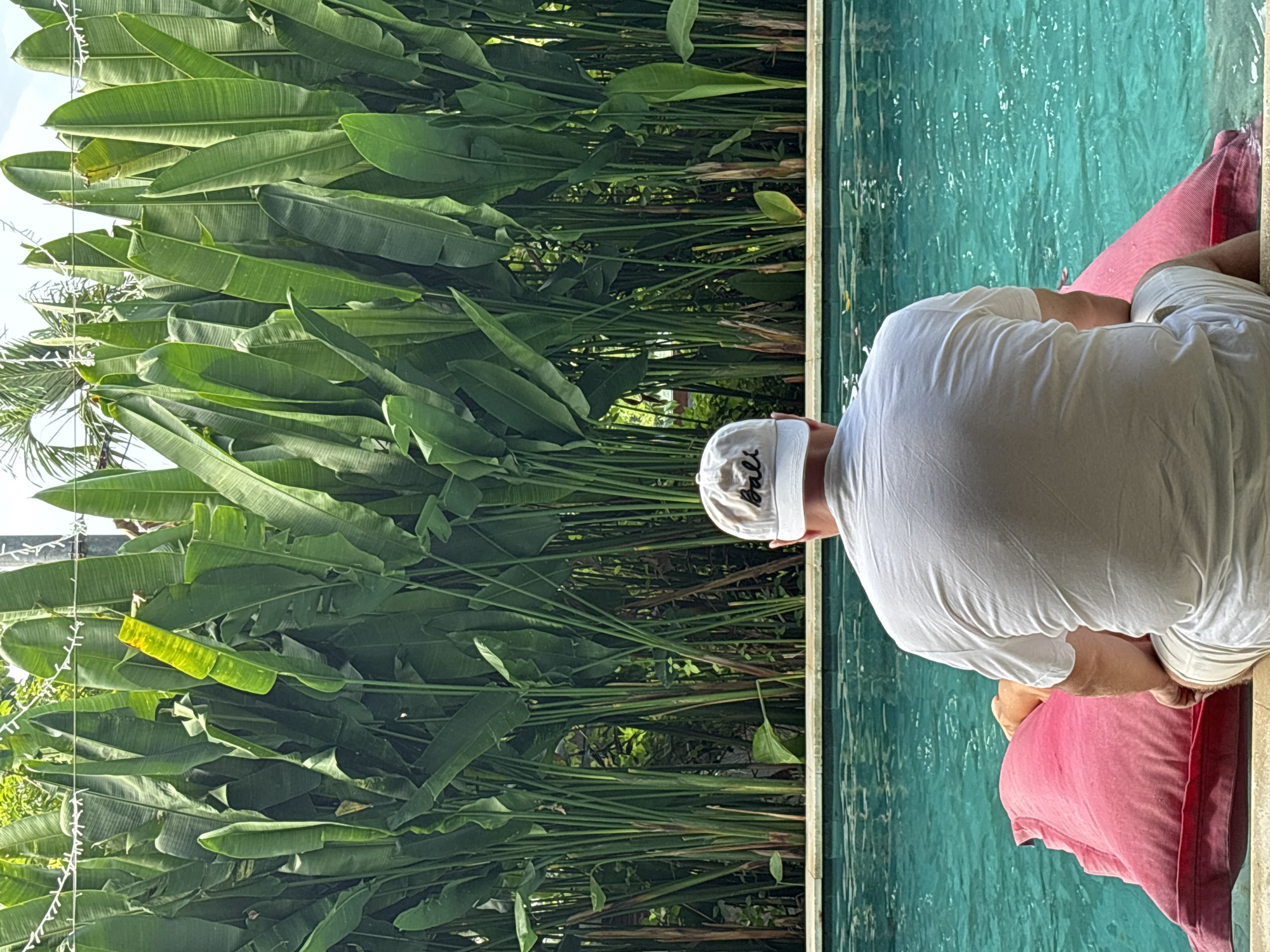 A person wearing a white t-shirt and white baseball cap is seen from behind, sitting on a pink towel next to a swimming pool with turquoise-colored water. Lush green foliage dominates the background, obscuring any buildings or structures. The location is Kuta Selatan, Indonesia.