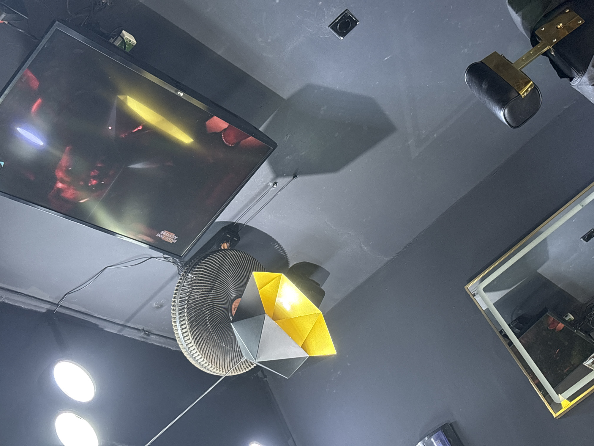 The image shows a low-angle, indoor shot from a location in Lugbe, Nigeria. The ceiling is a dark gray. Mounted on the ceiling is a television, a ceiling fan with an attached geometrical light fixture of gray and yellow, a small electrical outlet, and a framed mirror. Visible also, is a purse hanging from the ceiling. There are three bright circular lights reflecting off of the dark surface below the fan.