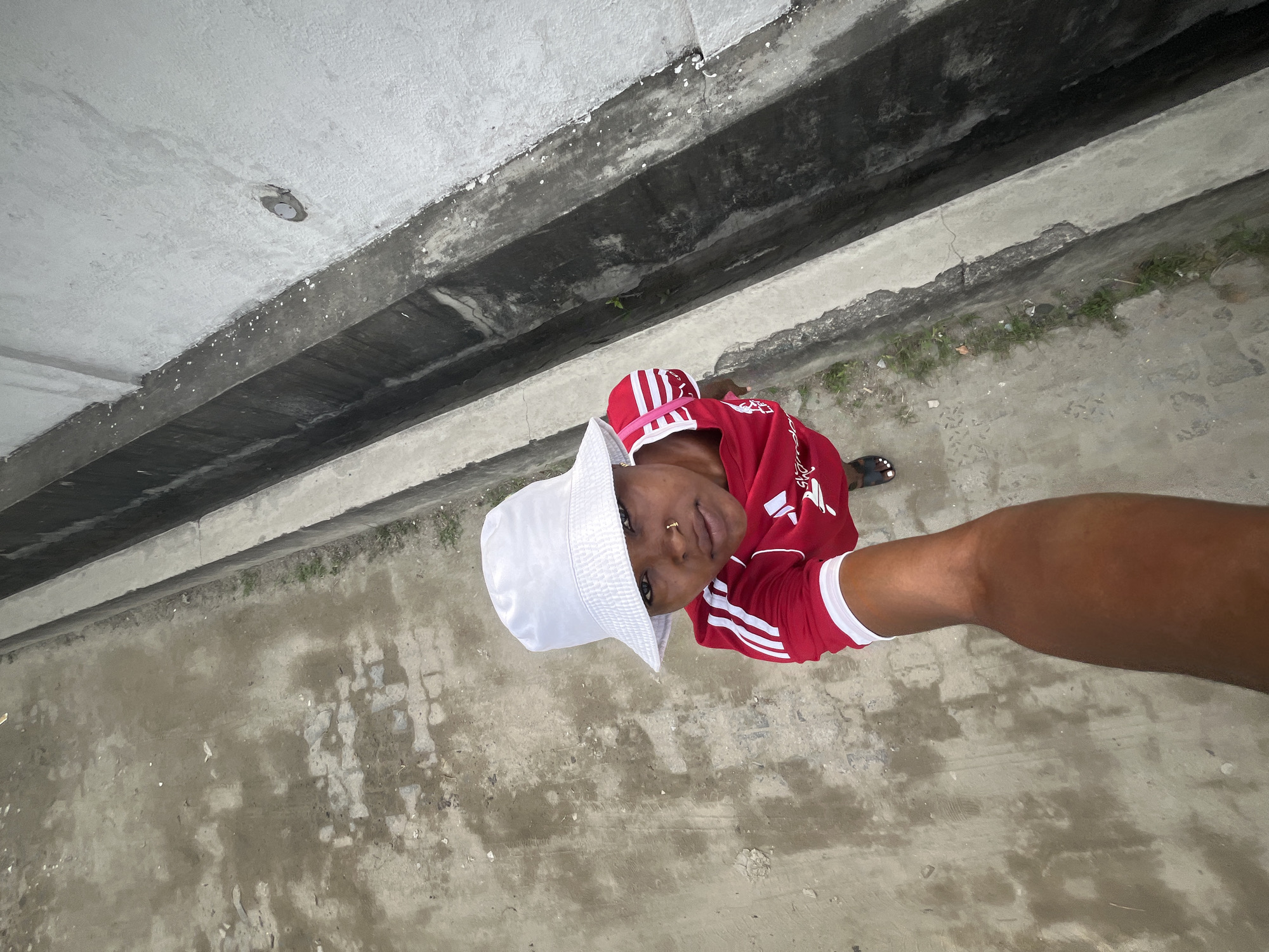 A high-angle, top-down shot captures a person standing outdoors, likely under an elevated concrete structure in Ado, Nigeria. The subject wears a white bucket hat, a red long-sleeved shirt with white stripes, and black sandals or slippers. They look directly upwards towards the camera, which appears to be held by another person, whose bare forearm is visible in the lower right of the frame, suggesting a selfie or close-up perspective.
The setting is characterized by a rough, light gray concrete or paved ground, interspersed with patches of dirt, small green weeds, and debris. Above the person, a massive concrete structure extends across the upper half of the image, featuring prominent dark, deep channels or grooves, and lighter concrete beams. This overhead structure suggests an underpass, a bridge, or the underside of a large building. The lighting indicates it is daytime, though the area beneath the structure is somewhat shaded. The red shirt displays white stripes on the sleeves and some indistinct white text or design, possibly in Arabic script, on the chest. The environment appears functional and weathered.