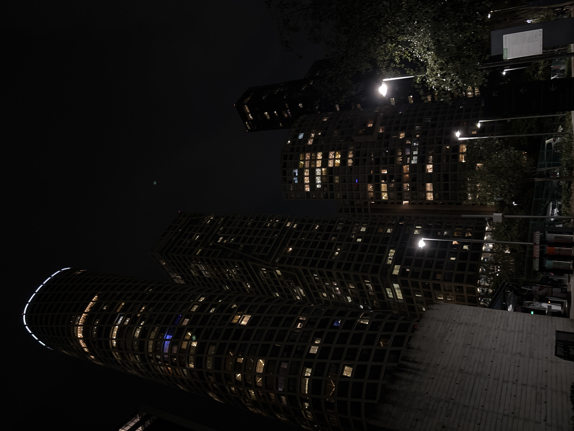 Here is a description of the image:
The image depicts a nighttime urban scene featuring modern architecture and street lighting. In the foreground and center are high-rise buildings with a distinctive grid-like facade, many windows lit from within, creating a pattern of light and shadow. The buildings have curved and angular forms, suggesting a contemporary design. Overhead street lights illuminate the area, casting light onto the surrounding structures and foliage. Dark silhouettes of trees and foliage are visible along the right side, obscuring portions of the buildings. There are signs and street furniture also visible along the side. The sky is dark and mostly featureless, contributing to the nighttime atmosphere of the city.