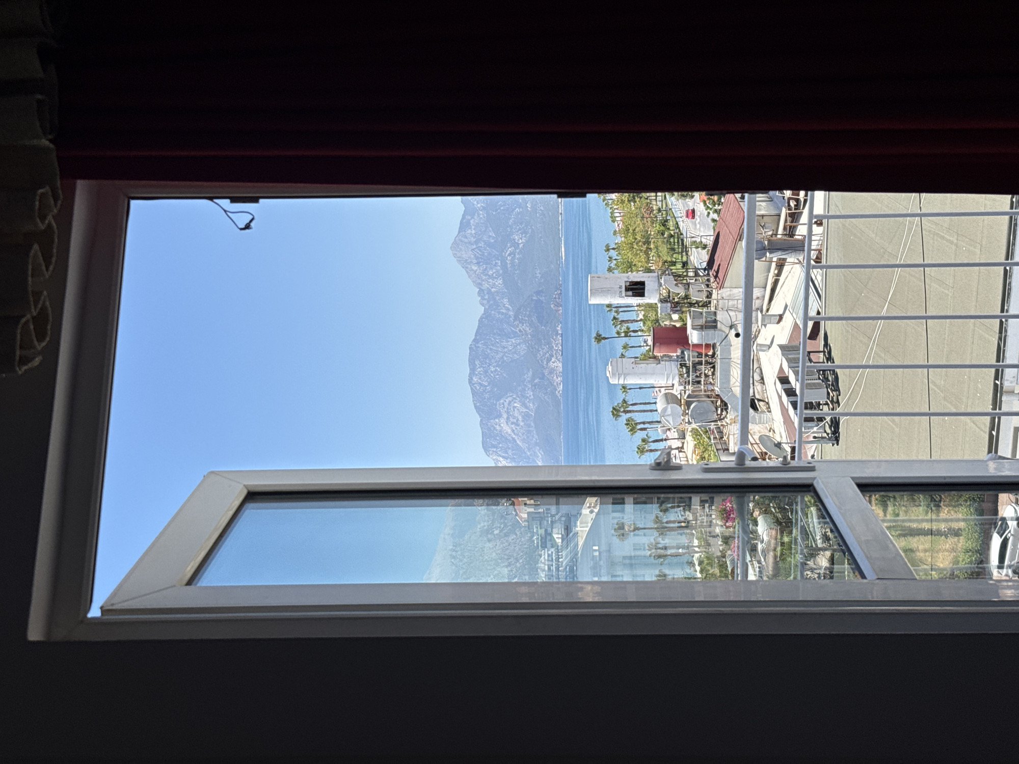 The image shows a view from a partially opened window looking out over a coastal city and the sea. In the foreground, the window frame and a dark red curtain are visible. Beyond the window, a bright blue sky dominates the upper half of the image.
The middle ground reveals a city scene with buildings, palm trees, and rooftops with air conditioning units and water heaters. A white metal balcony railing runs across the bottom of the visible outdoor space. A road with a red car can be seen in the distance, bordered by more trees.
The background features a serene blue sea meeting a range of large, mountainous islands. The overall scene suggests a warm, sunny day in a resort area, likely in Konyaaltı, Turkey, known for its beautiful coastline and mountainous backdrop.