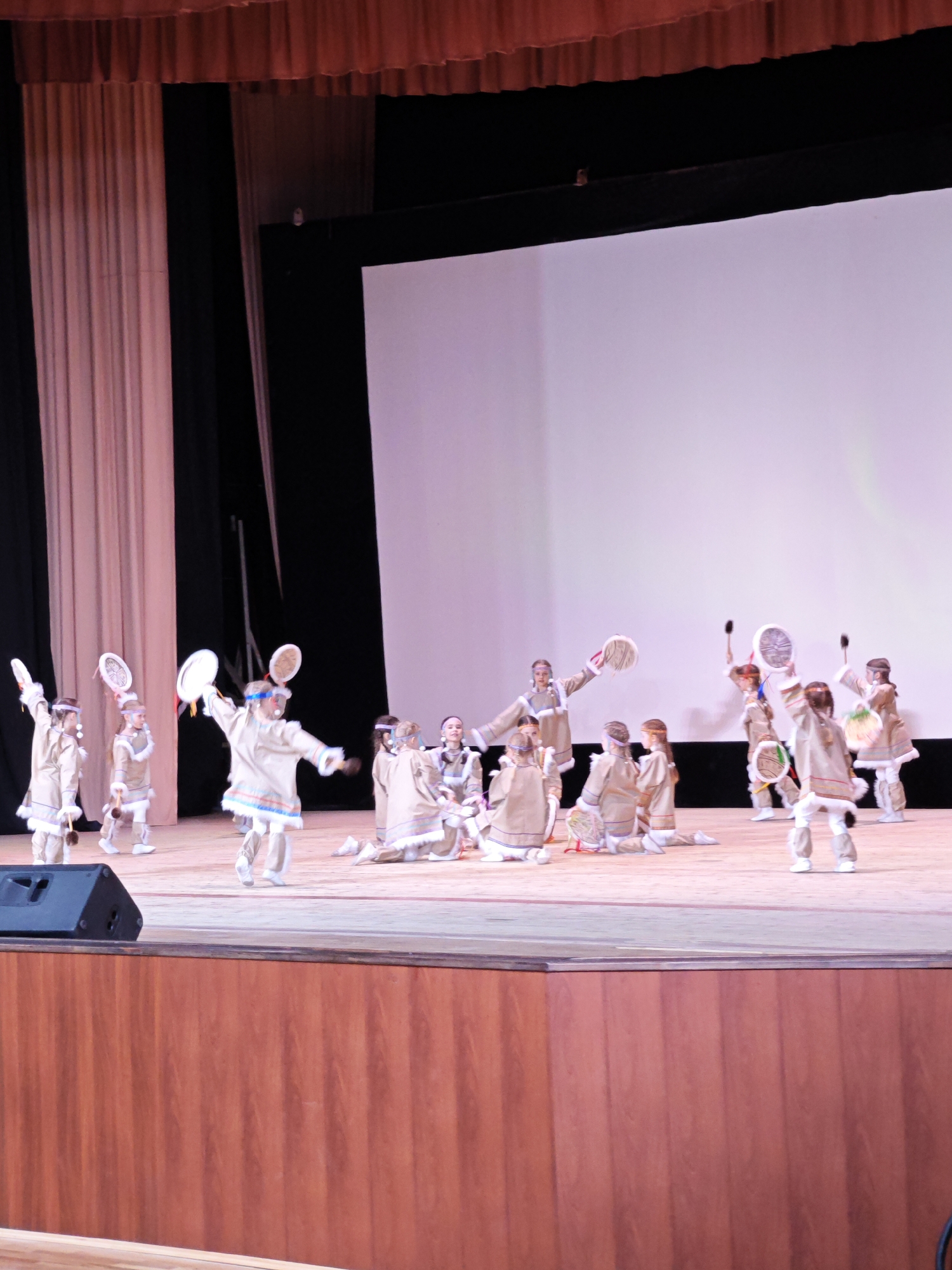 A group of children is performing on a stage in what appears to be a cultural performance. The stage has a wooden facade and is covered with curtains. A large white screen serves as a backdrop. The performers are dressed in similar light-colored costumes with fur trim and hold tambourines. The performance appears to be taking place indoors. The location is Марковский, Russia.