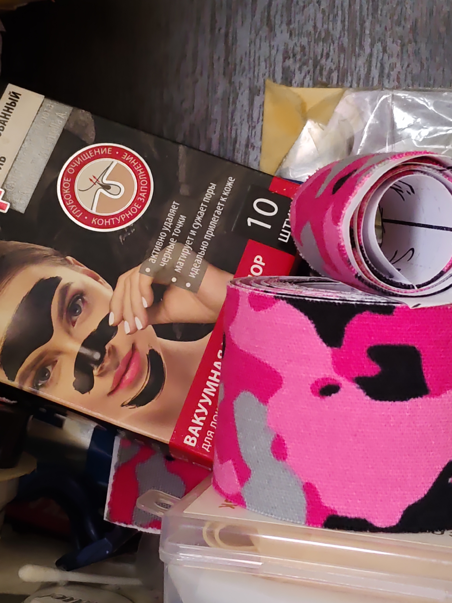 The image depicts a close-up view of a chaotic collection of beauty products and cosmetic items, likely stored in a drawer or box. In the foreground, a roll of pink and black camouflage-patterned fabric or tape is prominently displayed, with another similar roll partially visible behind it. To the left, a box for facial pore strips is visible, featuring an image of a woman with black pore strips applied to her face. Text in Russian is present on the box, indicating it is for "deep cleansing" and "contouring." Behind these items, other objects and packaging are jumbled together, suggesting a disorganized storage area. The overall impression is one of personal grooming supplies in disarray. The image does not provide any direct visual clues to its location within Rostov-on-Don, Russia, or any specific environmental details that would indicate an urban setting.