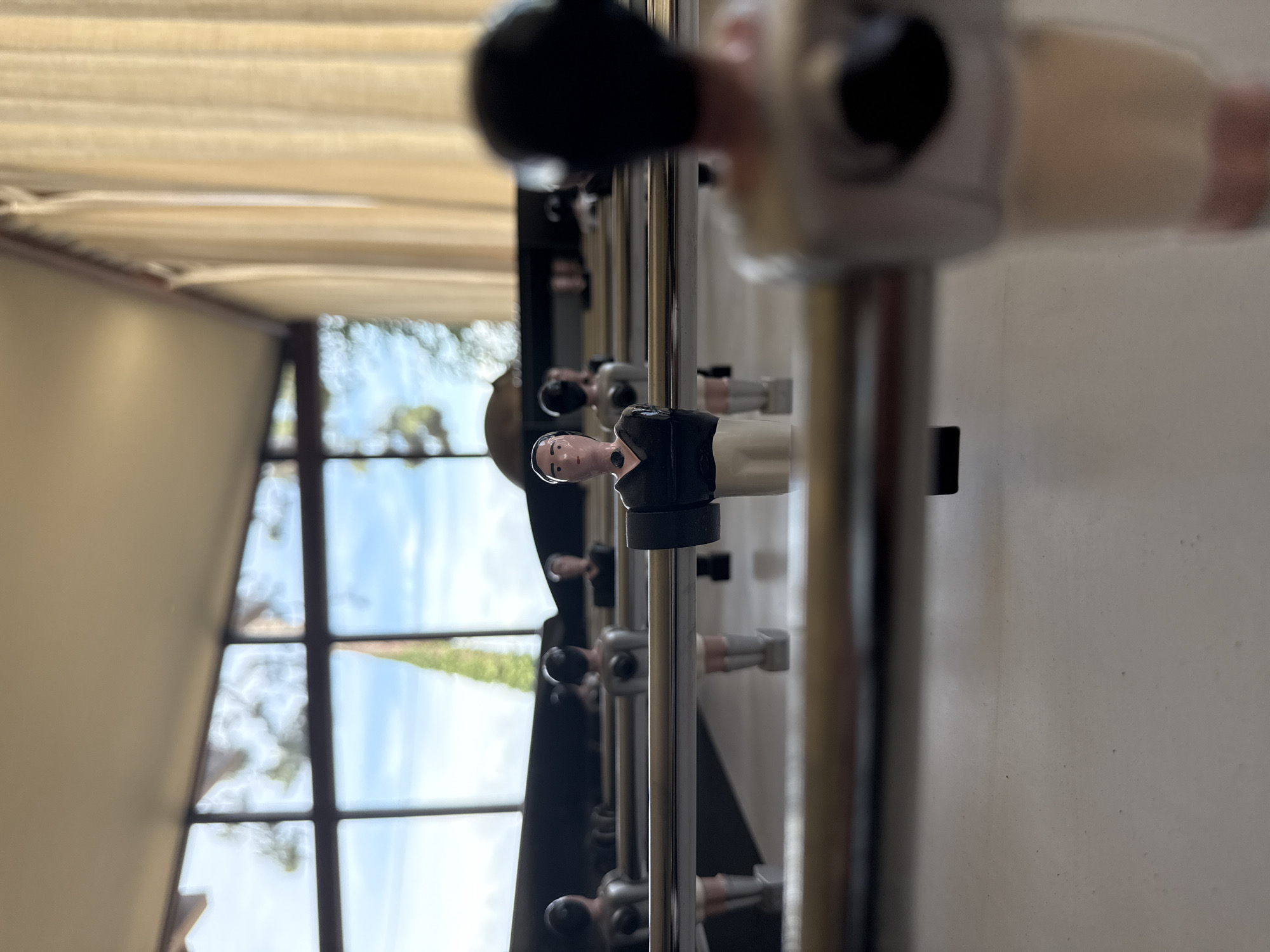 The image presents a close-up, low-angle perspective of a foosball table, initially rotated 90 degrees clockwise. When viewed correctly, a row of foosball player figures on their metal rods extends horizontally across the frame. The figure in the immediate foreground, dressed in a black top and white bottom, is sharply in focus, facing towards the left. Subsequent player figures and rods further down the table are progressively blurred.
The background reveals a large window with dark frames, offering a glimpse of the outdoors. Through the window, a bright blue sky with scattered white clouds is visible, along with blurred green foliage, indicative of a pleasant natural landscape. This natural light streaming in suggests the time of day is daytime.
Above the foosball table, a light-colored ceiling with horizontal blinds or slats is visible, also out of focus. A plain, light-colored wall forms the right side of the indoor setting. No specific activity or game play is depicted, only the static setup of the foosball table. There is no visible text in the image. The scene conveys a relaxed, recreational atmosphere within an indoor setting, likely in Lège-Cap-Ferret, France.