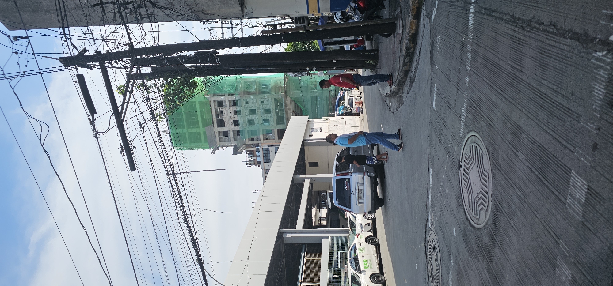 In Quezon City, Philippines, a street scene unfolds with a partially constructed building covered in green netting dominating the background. Overhead, a dense network of electrical wires crisscrosses the sky, supported by multiple utility poles. In the foreground, a grey asphalt road displays circular manhole covers with intricate patterns and painted white lines indicating traffic lanes. To the left, a white sedan with a "DRB" taxi marking is parked. Further down the street, a silver minivan, identified by its license plate "NAT 2253", is positioned on the road. Two men are engaged in conversation: one, wearing a blue t-shirt and jeans, stands on the road facing another man in a red t-shirt and jeans who is on the sidewalk. Another individual, a young male in a black t-shirt and patterned shorts, stands near the minivan. The environment appears to be a busy urban area, with evidence of ongoing construction and active transportation infrastructure.