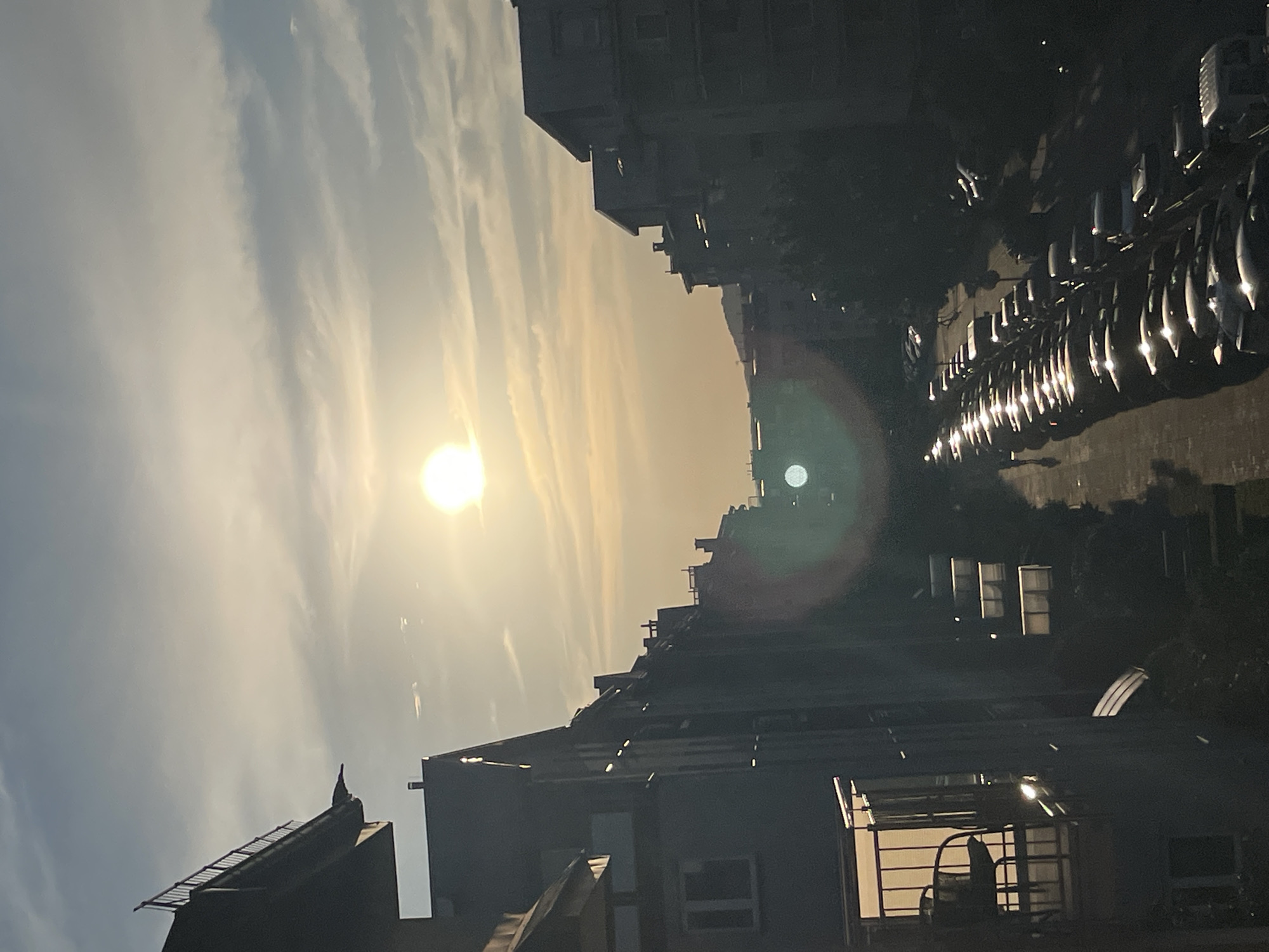 The image depicts an outdoor scene at sunset. A bright sun is visible in the sky, casting a warm glow and creating lens flare effects. The sky is partially covered with wispy clouds. In the foreground and mid-ground, there are several buildings, appearing to be apartment or office blocks. The architecture is modern and somewhat dense.
To the right of the buildings, a line of parked cars is visible. Their headlights are on, suggesting it might be dusk or the cars are parked in a dimly lit area. The overall impression is an urban or suburban setting. The time of day is clearly sunset, indicated by the sun's position and the warm, golden light. The weather appears to be clear with some cloud cover. There are no people clearly visible in the image. The scene evokes a sense of calm and the end of the day. No text is visible in the image.