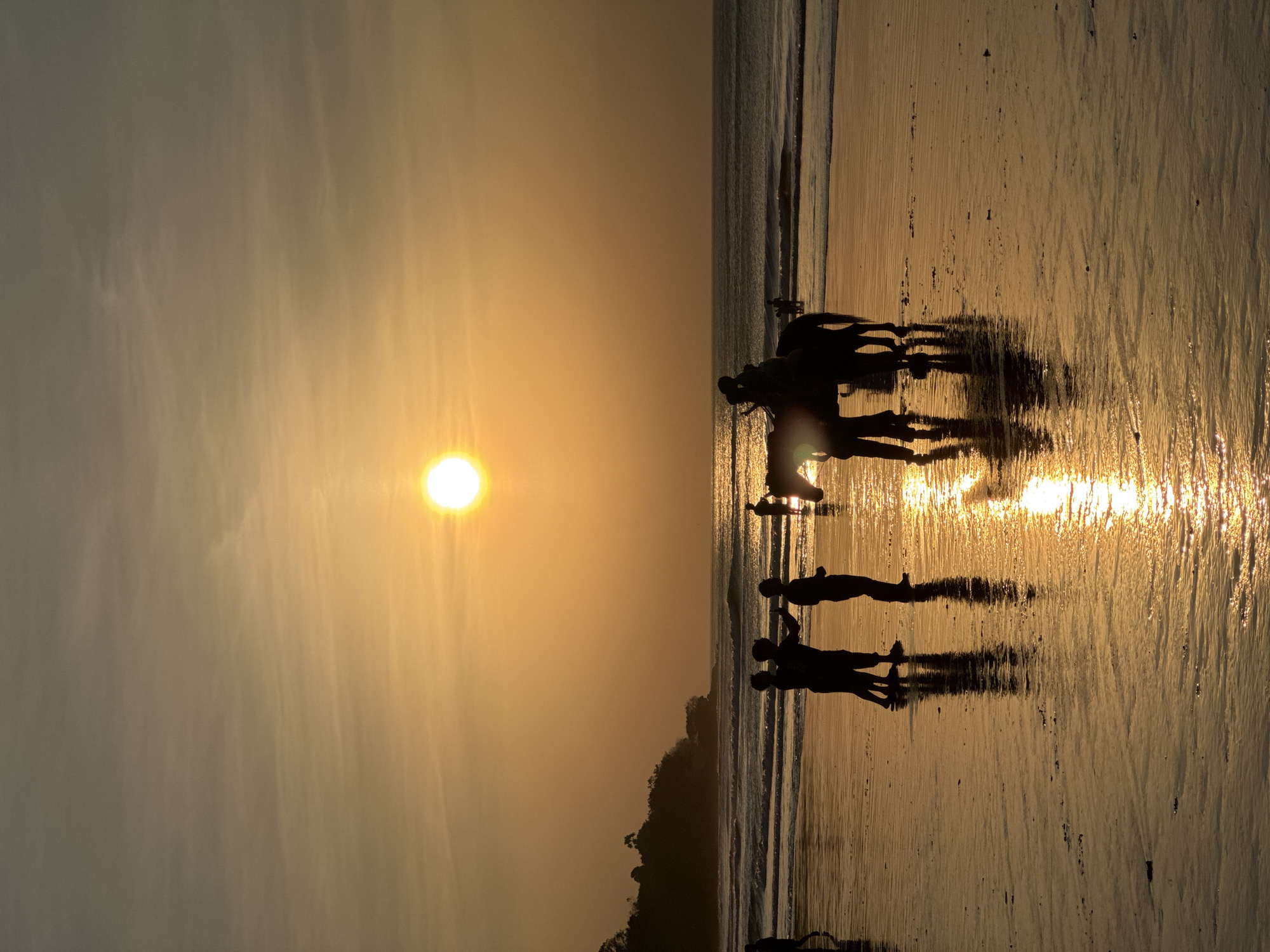 A vertical, high-angle, full shot captures a sunset on Kuta Selatan Beach, Indonesia. The sun, a bright orb, is positioned slightly left of center in the sky. Silhouetted figures are visible on the wet sand. In the foreground, three people stand together, their reflections appearing on the wet sand. To their right, a horse with two riders is also silhouetted, with its reflection visible. Further out, a lone figure stands in the shallow water, and more indistinct figures are visible further down the beach. Waves roll onto the shore in the midground, and a tree-covered hill forms the background on the left. The wet sand reflects the golden light of the sunset, creating a shimmering surface. The sky is a gradient of orange and yellow, with faint clouds visible.