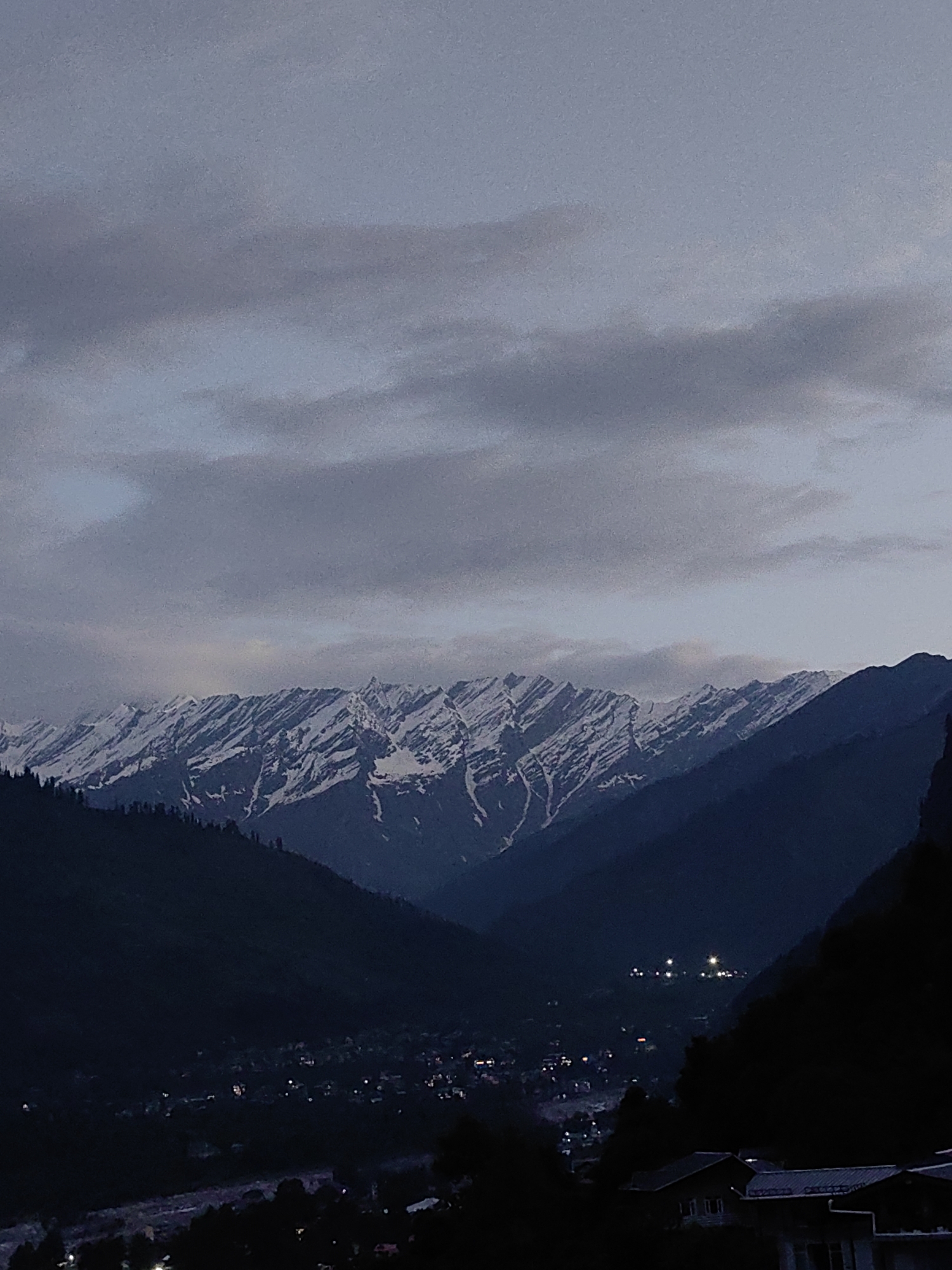 The image depicts a mountain range with snow-capped peaks, likely the Himalayas, under a cloudy twilight sky. In the foreground, dark, tree-covered slopes descend towards a valley. A small town or village is visible in the valley, illuminated by scattered lights, suggesting it is nighttime or dusk. The architecture of the buildings in the lower right portion of the image appears to be traditional, with sloping roofs. The scene is serene and atmospheric, conveying a sense of natural grandeur and the presence of a small human settlement nestled within the mountains.