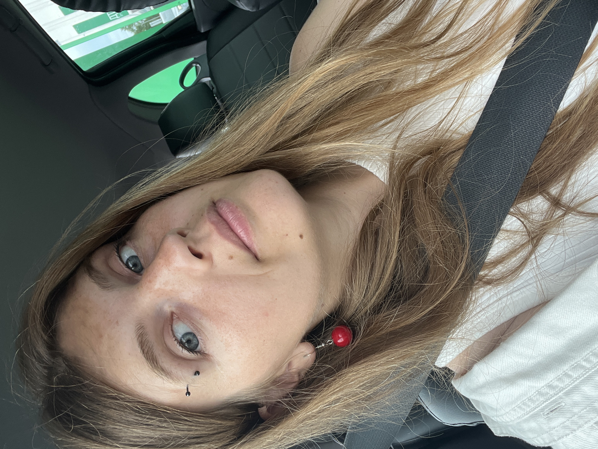 A woman with blonde hair and blue eyes is sitting in the driver's seat of a car. She is wearing a white top and a seatbelt. She has eyebrow piercings and a red earring. The car appears to be at a gas station, with a green and white sign visible through the window. The location is Шчомысліцкі сельскі Савет, Belarus.
