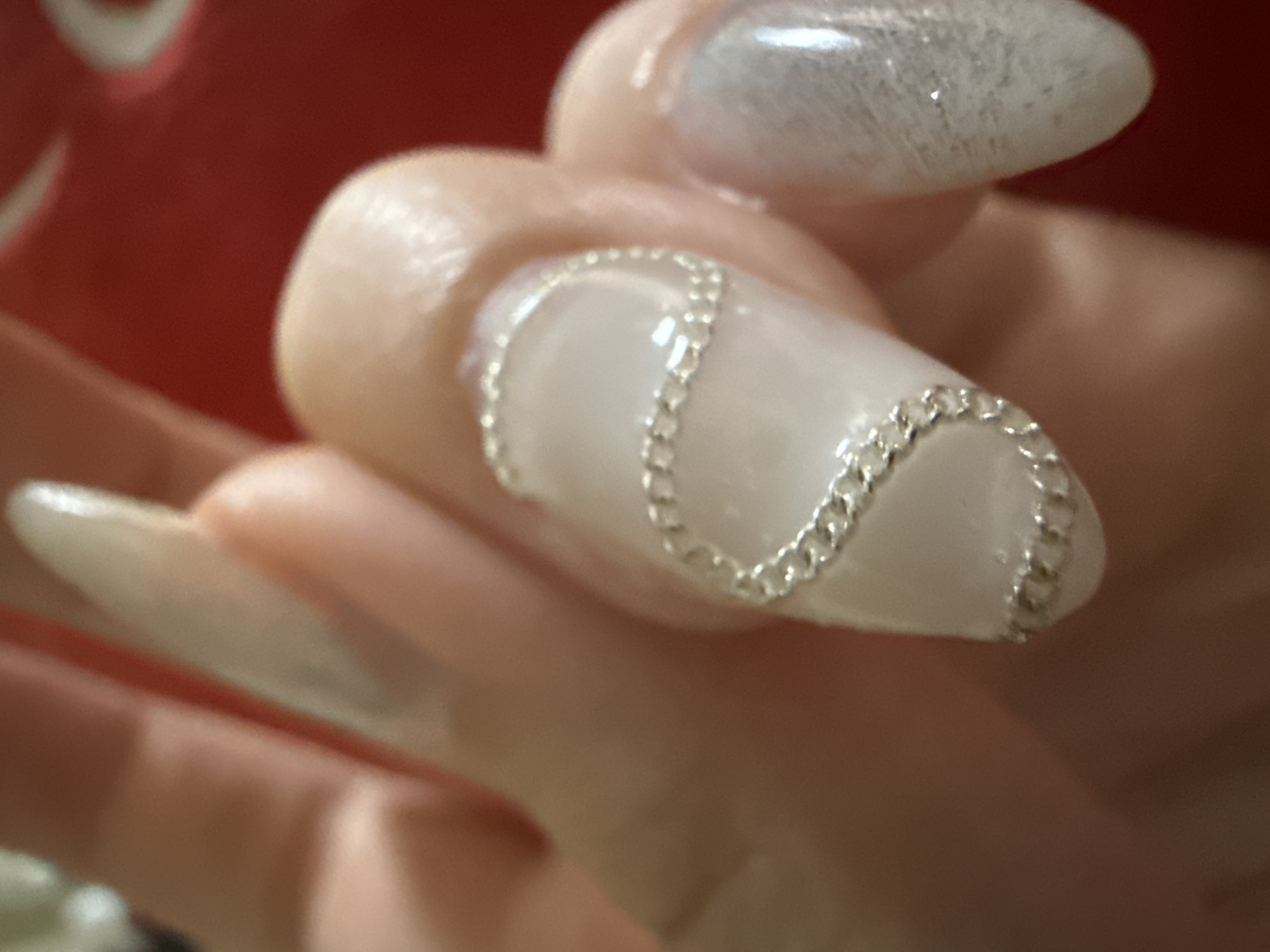 The image shows a close-up of a hand with long, pointed nails. One nail has a white base with a silver chain-like design curving across it. Another nail, slightly out of focus, appears to have a shimmery white design. The hand is holding something red in the background.