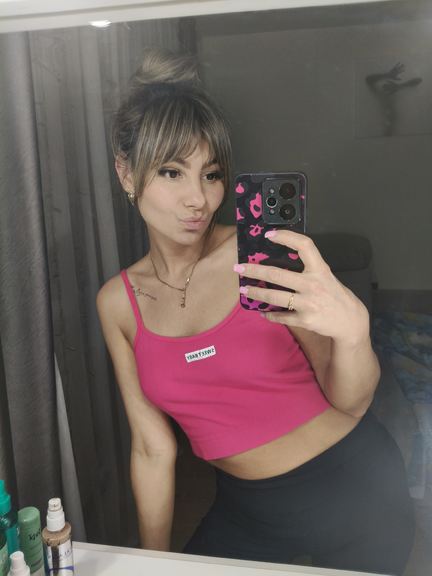 A young woman is depicted in a mirror selfie, posing with slightly pouted lips and looking towards her smartphone. She has light brown hair with blonde highlights, styled in a messy top bun with bangs. She wears delicate earrings, including a triangular dangle on her left ear, and a gold necklace with a pendant. Her right hand, with pink painted nails and a gold ring, holds a smartphone featuring a black case with pink leopard print.
The woman is dressed in a bright pink ribbed crop top, which has a white label on the chest displaying the text "SWEET BABY" (reversed in the mirror). She is also wearing black leggings. A faint tattoo of script is visible on her left collarbone area.
The background, reflected in the mirror, shows dark grey curtains on the left. Above her right shoulder, a wall decoration resembling a silhouette of a person with outstretched arms is visible. To the right, part of a dark sofa or headboard and a blue and white patterned blanket can be seen. On a counter or vanity in the bottom left, several cosmetic bottles are present, including a green spray bottle, another green pump bottle, and a beige pump bottle with the reversed text "COLLAGEN" and "LOREAL" visible on its label. The scene suggests an indoor setting, likely a private room.
