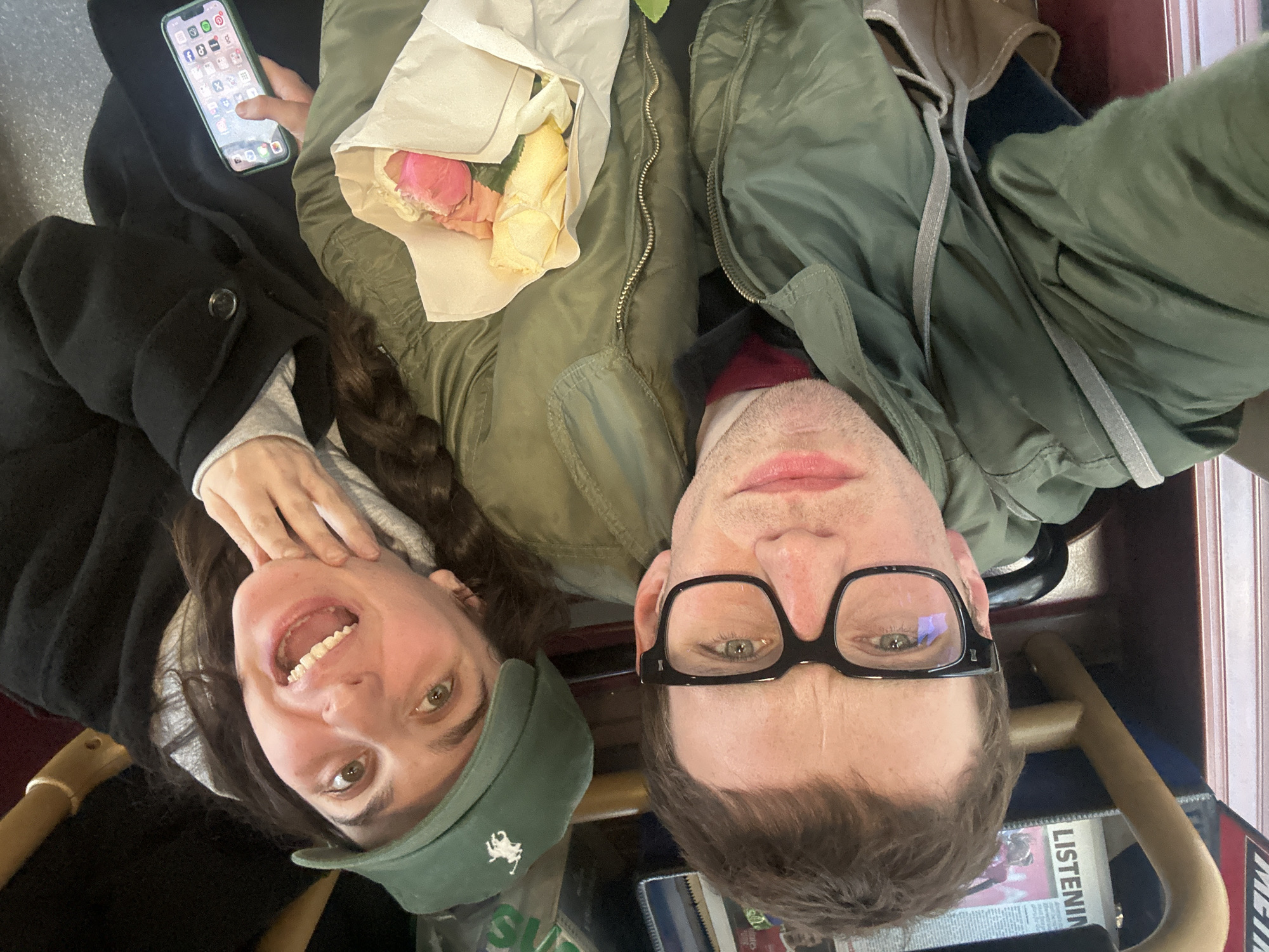 Two individuals are angled together in what appears to be a public transportation setting, likely a train or bus due to the presence of overhead railing and seating. One person, with a green baseball cap, smiles directly at the camera and is wearing a black coat over a grey sweatshirt. They have long, wavy hair and are holding their face with one hand. The other person, next to them, wears eyeglasses and a green bomber jacket, and is looking at the camera. Between them is a paper bag containing flowers, specifically pink and yellow roses. One person has a mobile phone in hand. The location is likely London, United Kingdom, given the presence of the Metro newspaper and the style of seating.