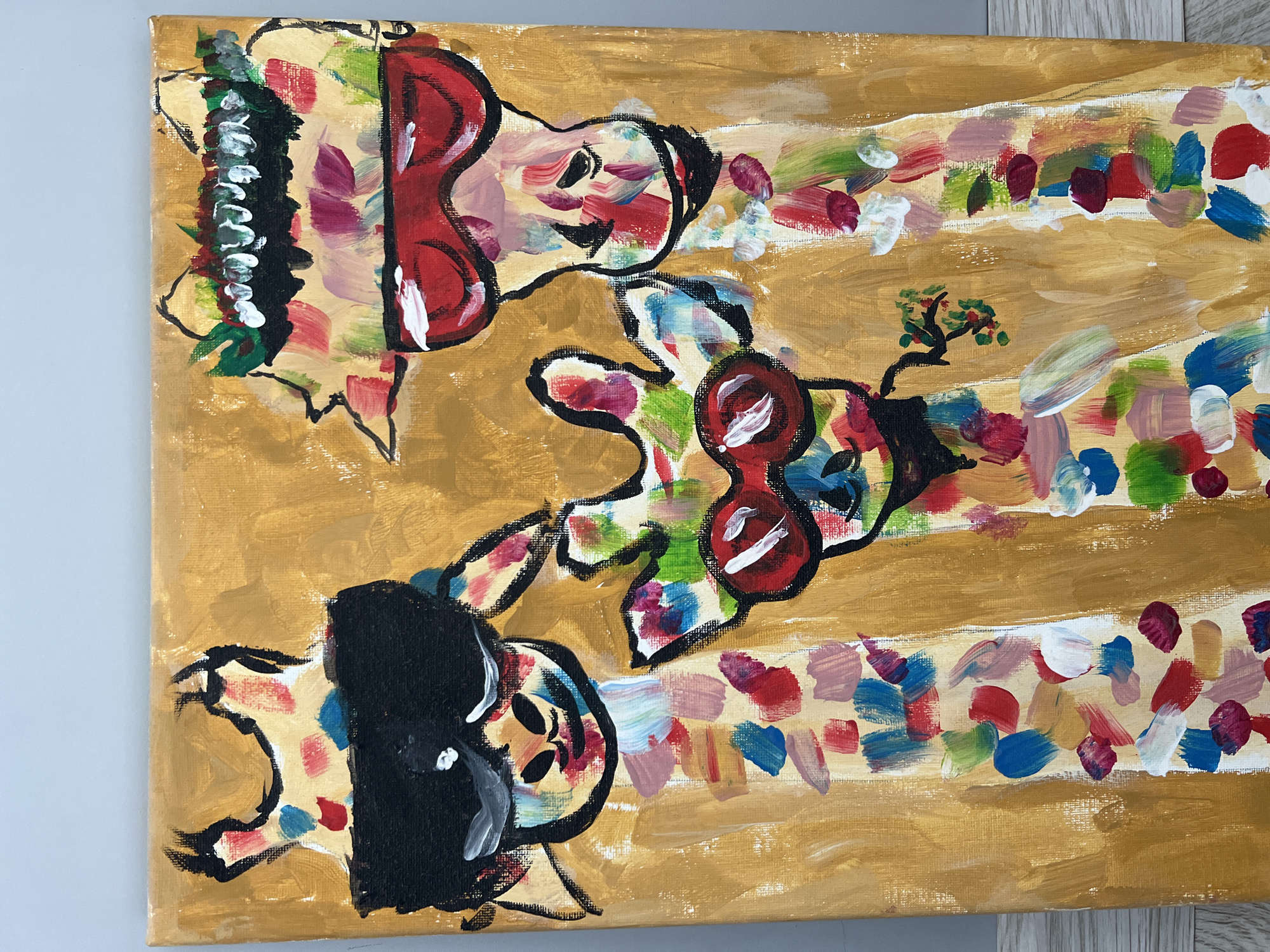 The image shows a canvas painting. The background is a mottled gold color with faint horizontal stripes. There are three abstract figures painted on the canvas, each appearing to be portraits. The top left figure has red hair and a black collar, while the figure on the bottom left has black hair and a white collar. The central figure appears to have a potted plant growing from the top of their head. The figures are outlined in black and have splashes of color within the outlines. The location is unknown.