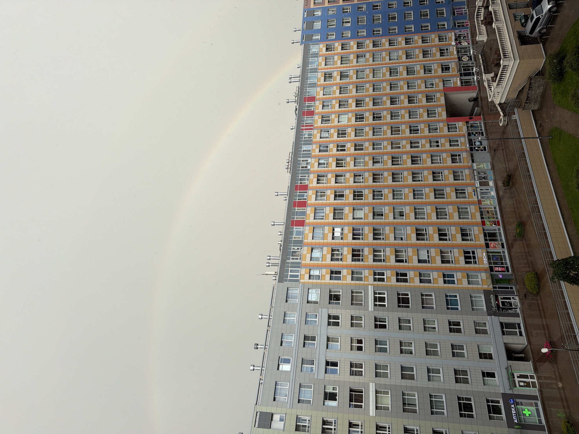 The image shows a wide, panoramic view of a modern, multi-story residential building complex under a cloudy sky with a faint rainbow visible. The building has a varied facade, with sections in gray, orange and yellow checkered patterns, and blue. The lower floors feature commercial establishments, including "АПТЕКА" (Apteka, meaning Pharmacy), "OZON", and other shops. A street is visible in front of the shops, with some scattered people and a car parked in the background. The ground appears wet, suggesting recent rain. The architecture is contemporary and urban.
