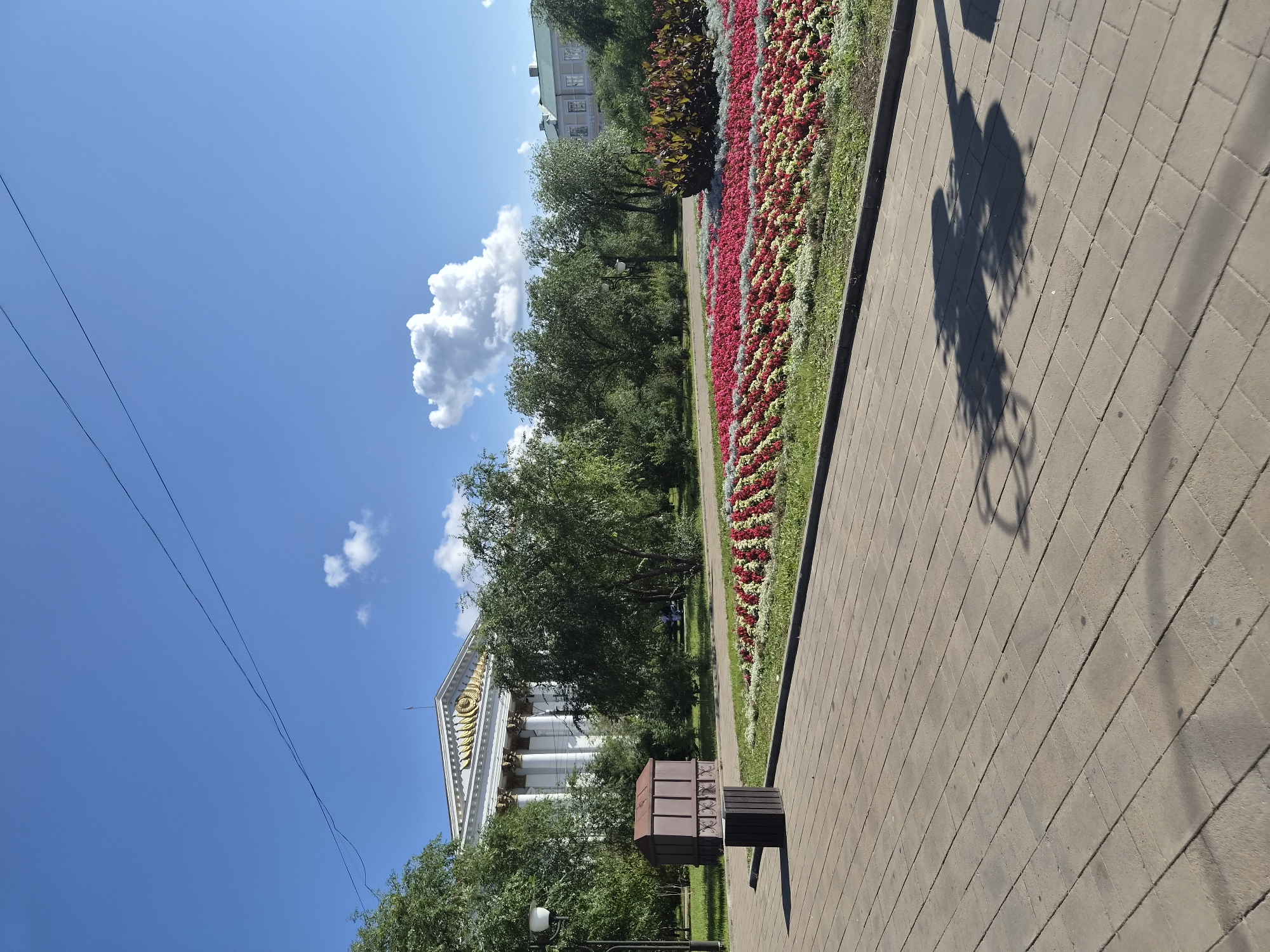 In an outdoor urban setting under a clear blue sky with scattered clouds, a paved walkway in the foreground leads toward a lush park area. The walkway is constructed of rectangular stone pavers. To the left of the walkway, there is a brown trash receptacle. Beyond the walkway, a vibrant flowerbed features rows of red, white, and pink flowers, bordered by greenery. Behind the flowerbed, a line of trees separates the park from a large, white building with classical architecture, characterized by its columns and ornate facade. To the right of the main building, a second building with a lighter facade and green roof is partially visible. The scene is bathed in sunlight, casting shadows from trees and a lamppost onto the walkway.