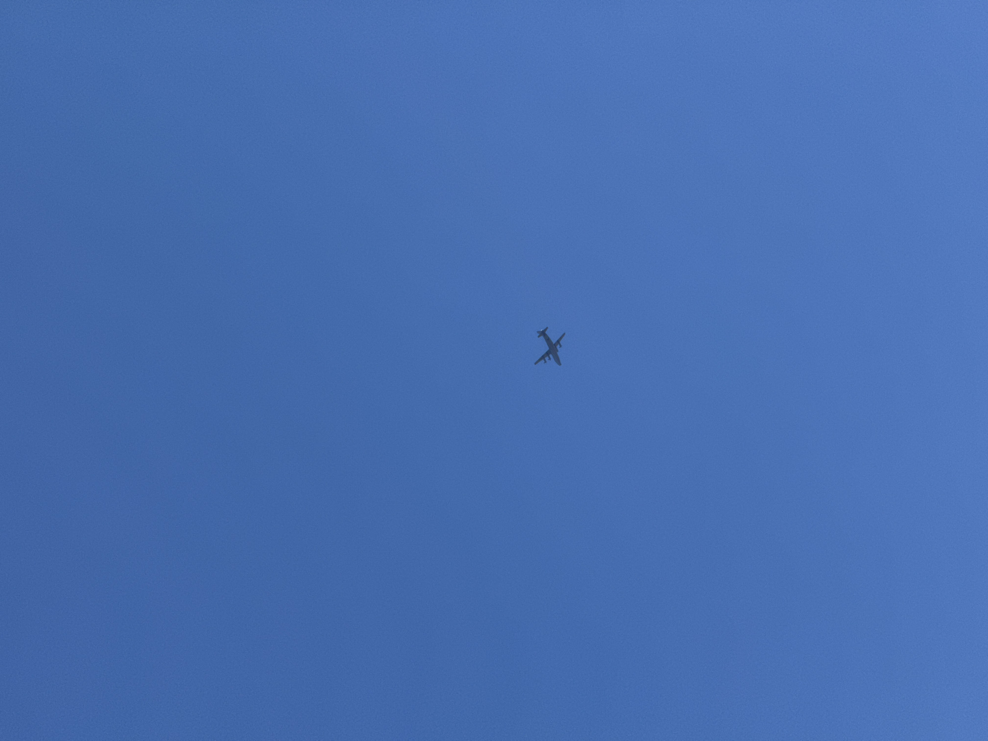 The image displays a clear, unblemished deep blue sky with a single airplane flying across it. The aircraft is positioned near the center of the frame, appearing as a dark silhouette against the bright sky, indicating it is likely at a high altitude.
The airplane is the sole subject, rendered small due to the distance. Its shape suggests a multi-engine propeller aircraft or a similar type with engines under the wings, and a distinct tail structure is visible. The wings are spread wide, and the body tapers towards the tail.
The scene is set against a vast, cloudless blue sky, suggesting excellent weather conditions during the daytime. There are no other visible objects, landmarks, or features, giving a sense of openness and isolation for the flying aircraft. No people are visible, nor is any text. The uniform blue of the sky is consistent across the entire image. The location context for this image is Лобаново, Russia.