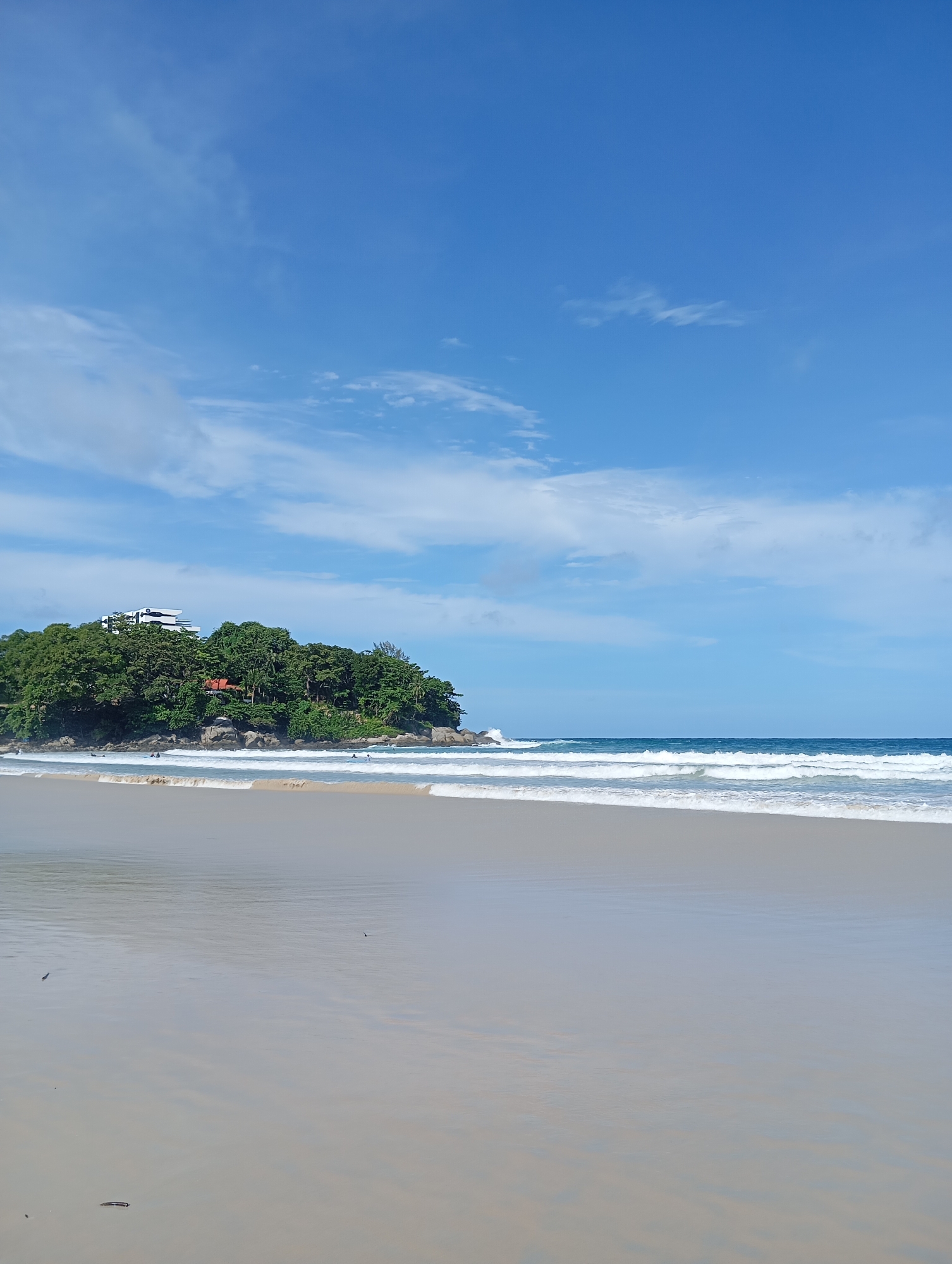 The image depicts a serene beach scene in Karon, Thailand. The foreground is dominated by a wide expanse of wet sand, reflecting the sky and subtly showing ripples from receding waves. The middle ground features the gentle, foamy surf of the ocean with several parallel waves breaking onto the shore. In the background, a lush green headland juts out into the sea, densely covered with trees. A modern, white building with a distinctive angular design is visible atop the headland, partially obscured by foliage. Scattered among the trees on the headland are hints of red-tiled roofs, suggesting smaller structures. Several small figures, presumably people, are visible in the water and on the distant edge of the beach, engaged in recreational activities. The sky above is a clear, bright blue, punctuated by scattered white clouds, creating a picturesque tropical atmosphere.