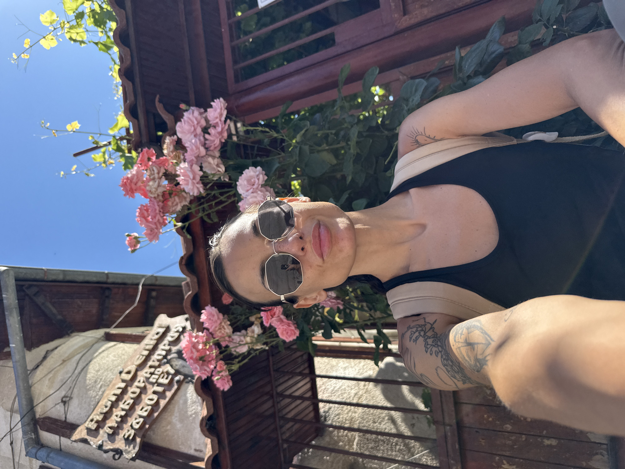 In Pleven, Bulgaria, a woman with dark hair and sunglasses poses for a selfie in front of a building with a sign written in Cyrillic. She is wearing a black tank top and a light-colored backpack. Pink roses are blooming on a vine that climbs the building. The sun is shining brightly, casting shadows on the stone wall and wooden gate behind her. A drainpipe runs down the side of the building.