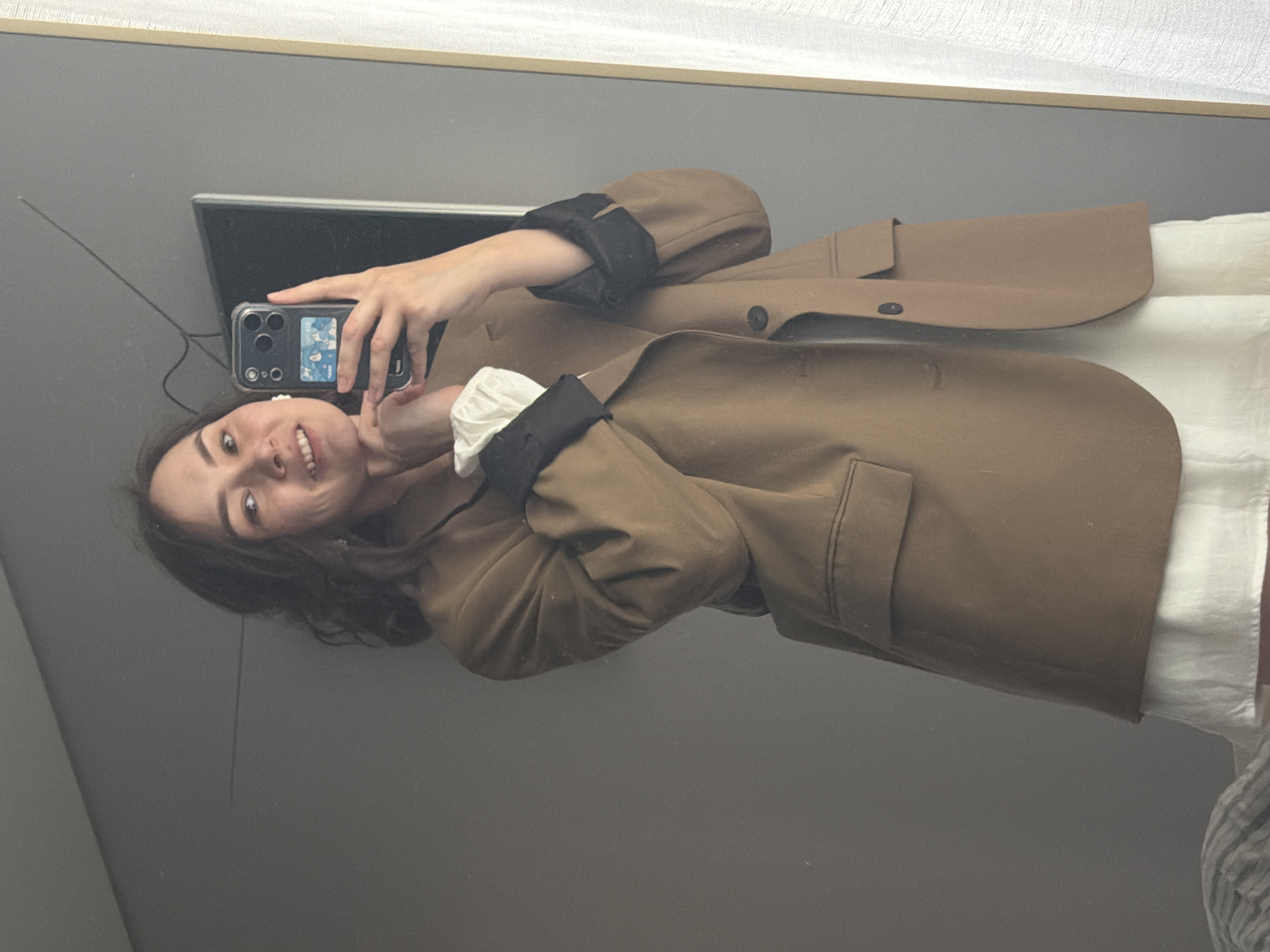 A young woman with dark wavy hair is taking a mirror selfie in an indoor setting. She is wearing an oversized tan blazer over a white dress. Her left hand is resting on her chin, and she is smiling at her reflection. She is holding a smartphone in her right hand, which is also visible in the reflection. The background appears to be a room with gray walls and a white curtain to the right. The image is a medium shot, focusing on the woman's upper body and face. There is no visible indication of the city of Chaikovsky, Russia in the image.