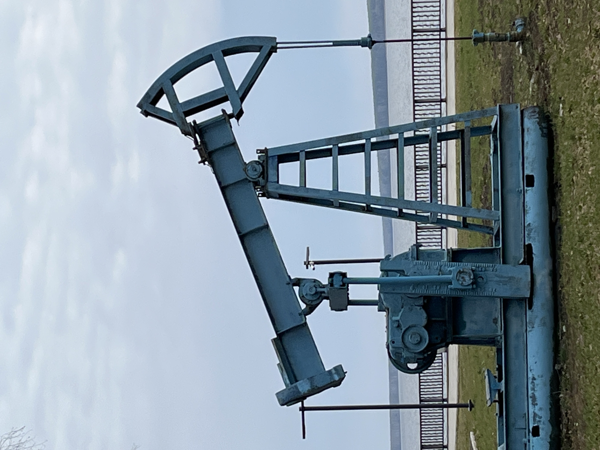 The image shows a blue-painted oil pump jack, situated outdoors near a body of water (potentially the Kama River) and a black metal railing. The pump jack is oriented vertically in the frame. There is some grass and vegetation in the background. The sky is overcast. Given the location, this is likely the oil pump jack memorial in Chaikovsky, Russia.