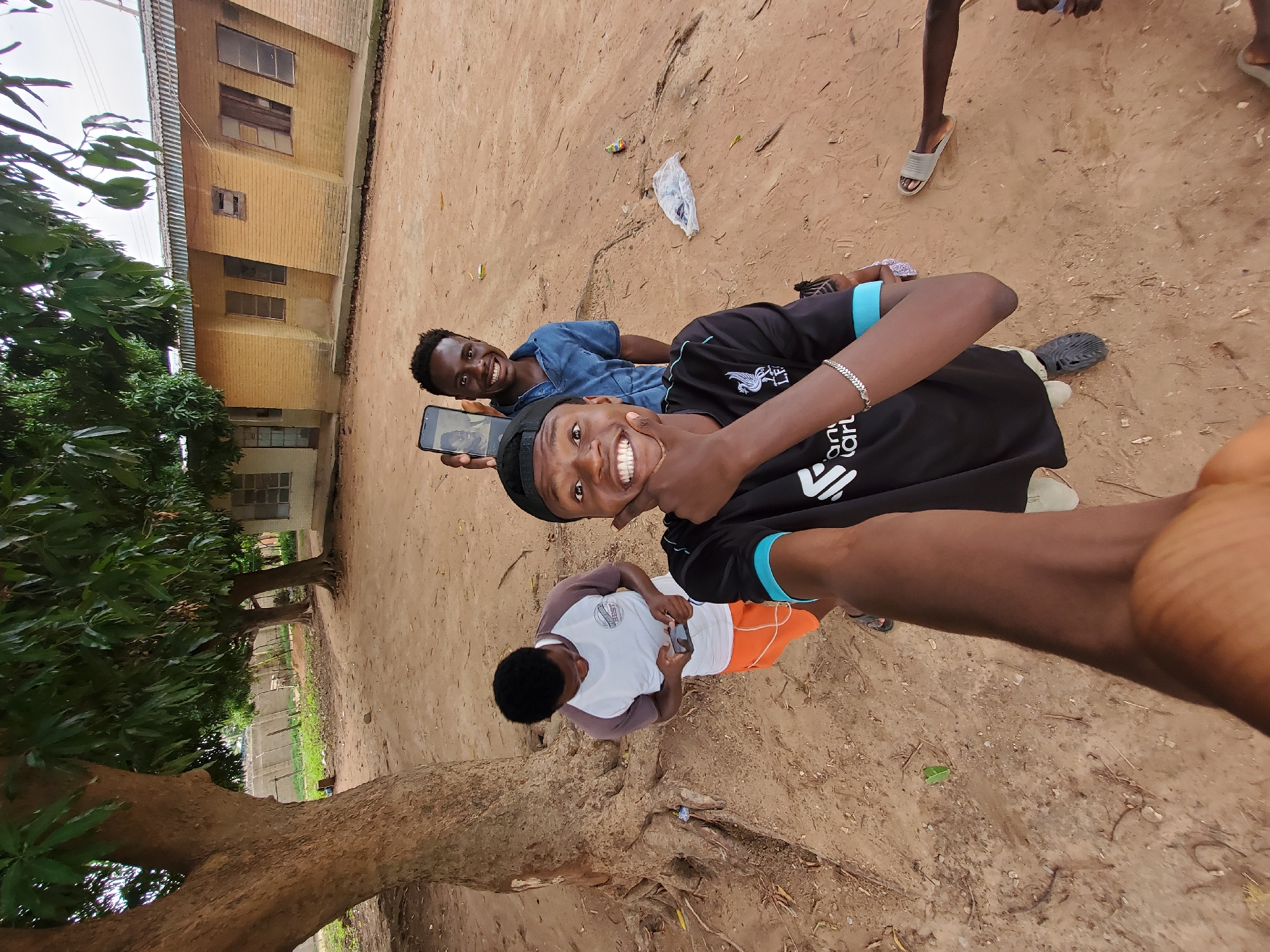 In Narayi, Nigeria, three young men are captured in a selfie. One man, positioned in the foreground and slightly to the right, holds his left hand to his chin, smiling widely. He wears a black t-shirt with a white logo on the left chest and a light blue collar. To his left, a second man, also smiling, stands behind a tree's trunk. He is dressed in a blue short-sleeved shirt and holds up a smartphone, displaying a picture of two individuals. To the left of the man with the smartphone, a third man is partially visible, wearing a white and brown t-shirt and orange shorts, looking down at a phone in his hands. The setting appears to be an outdoor area with a dirt ground, a large tree to the left, and a yellow brick building with corrugated metal roofing in the background. Several pieces of trash are scattered on the ground.