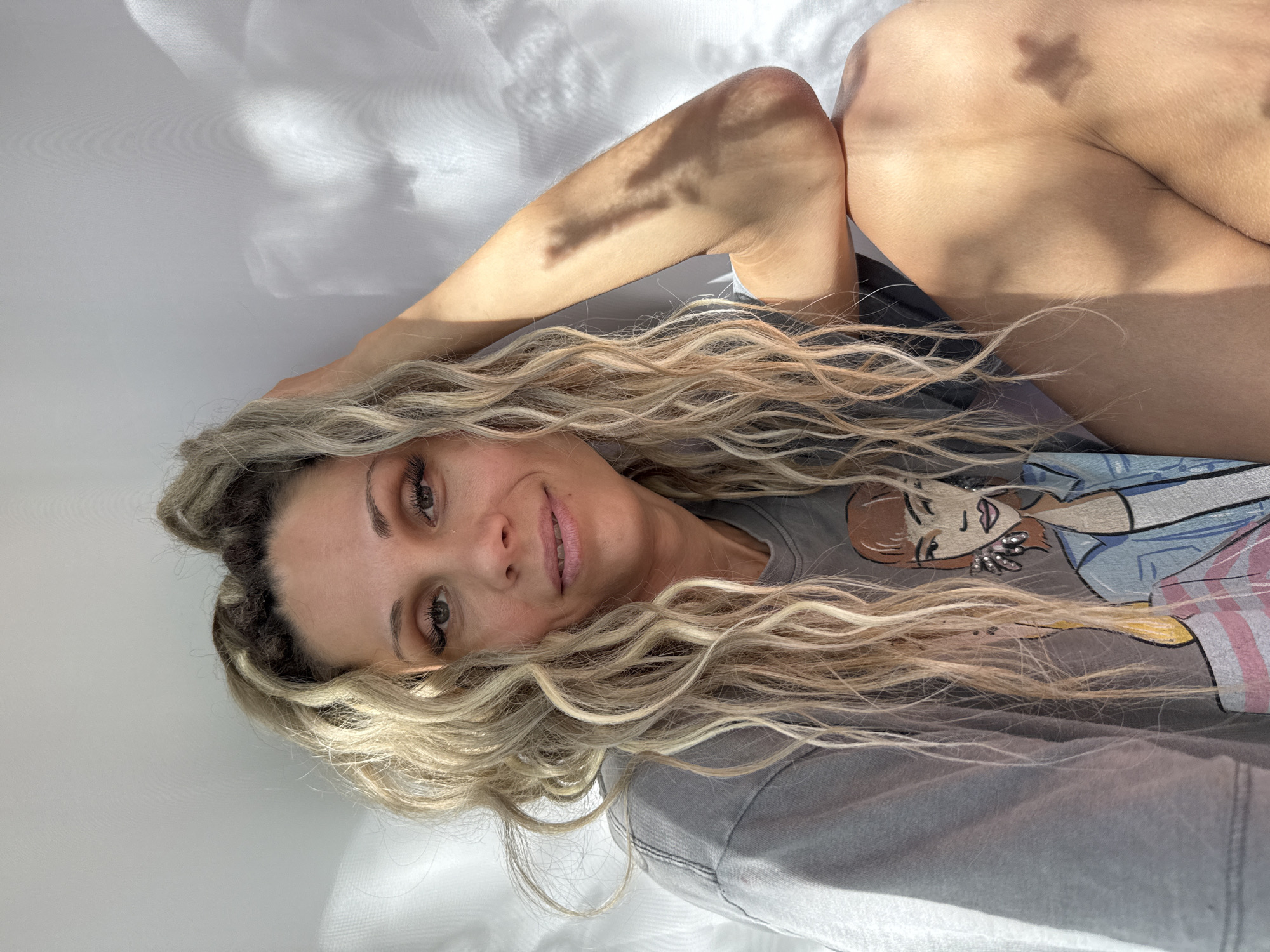 A woman with blonde, curly hair is sitting against a white wall. She is wearing a gray t-shirt with a cartoon graphic of a woman on it. The woman in the photo is looking at the camera and smiling slightly. Her left arm is bent, and her hand is resting on her head. Sunlight is casting shadows on the wall and the woman's arm and shoulder, creating a dappled effect. There are no specific details in the image that would indicate its location in Rusanovo, Russia.